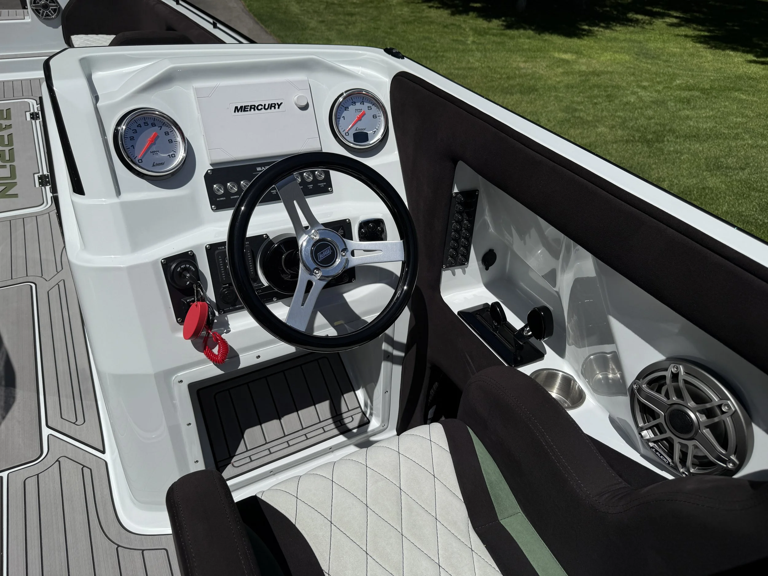 Interior view of a boat dashboard with white and black accents, two round gauges, a steering wheel with a Mercury logo, a panel of switches, a cup holder, and seating with a quilted pattern. Outside green grass is visible.