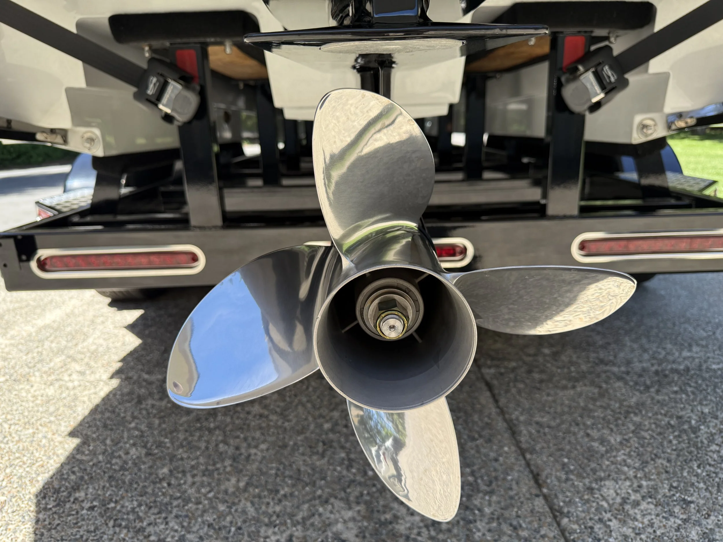 Close-up of a boat propeller with three shiny metal blades, mounted below a boat with a visible engine and black frame, on a paved surface with grass in the background.