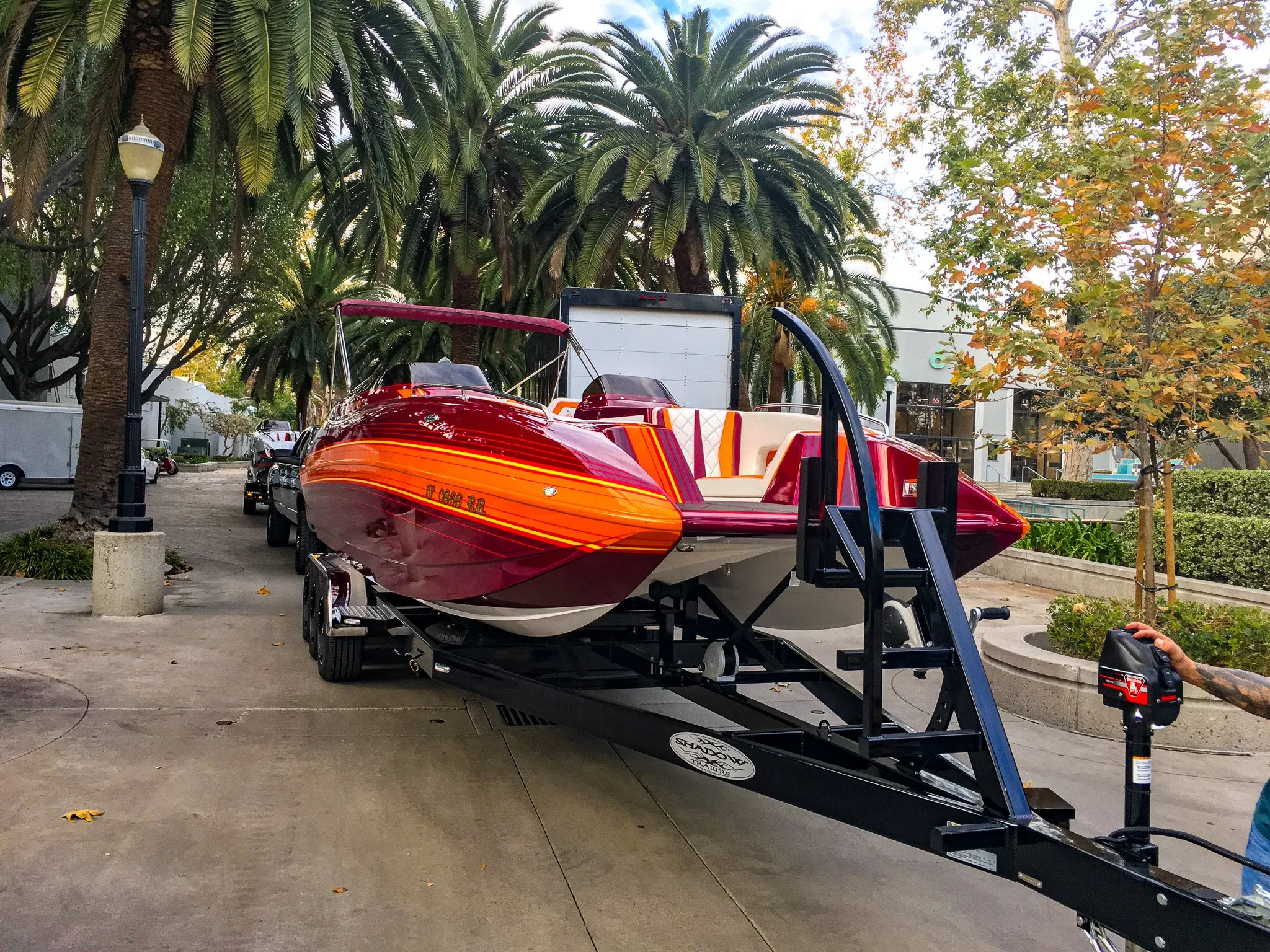 A red and orange speedboat on a trailer, parked in a lot with tall palm trees and deciduous trees with autumn leaves in the background.