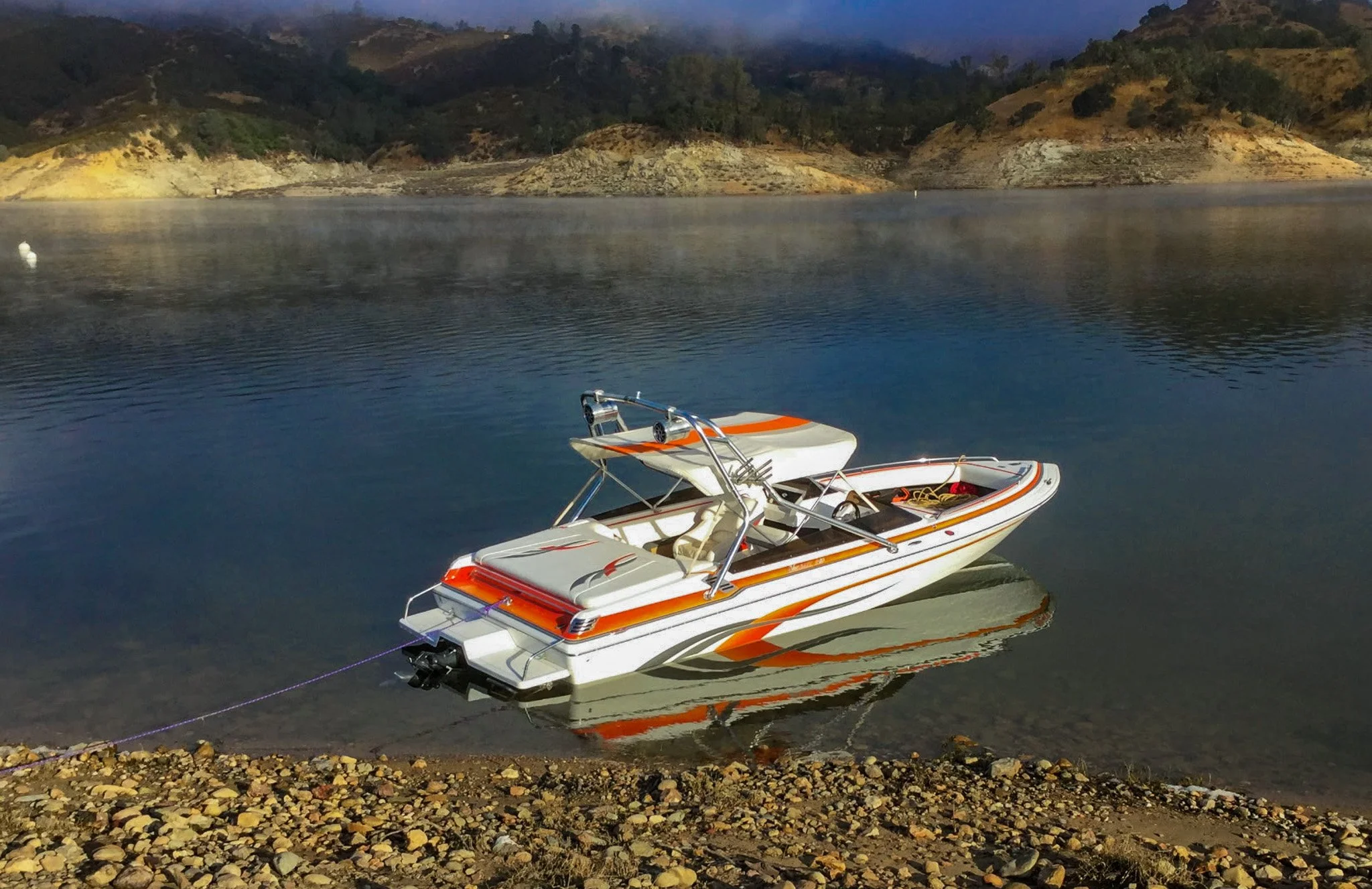 A speedboat with white, orange, and gray colors is docked near a rocky shoreline on a calm body of water, with distant green hills and mountains in the background during dusk.