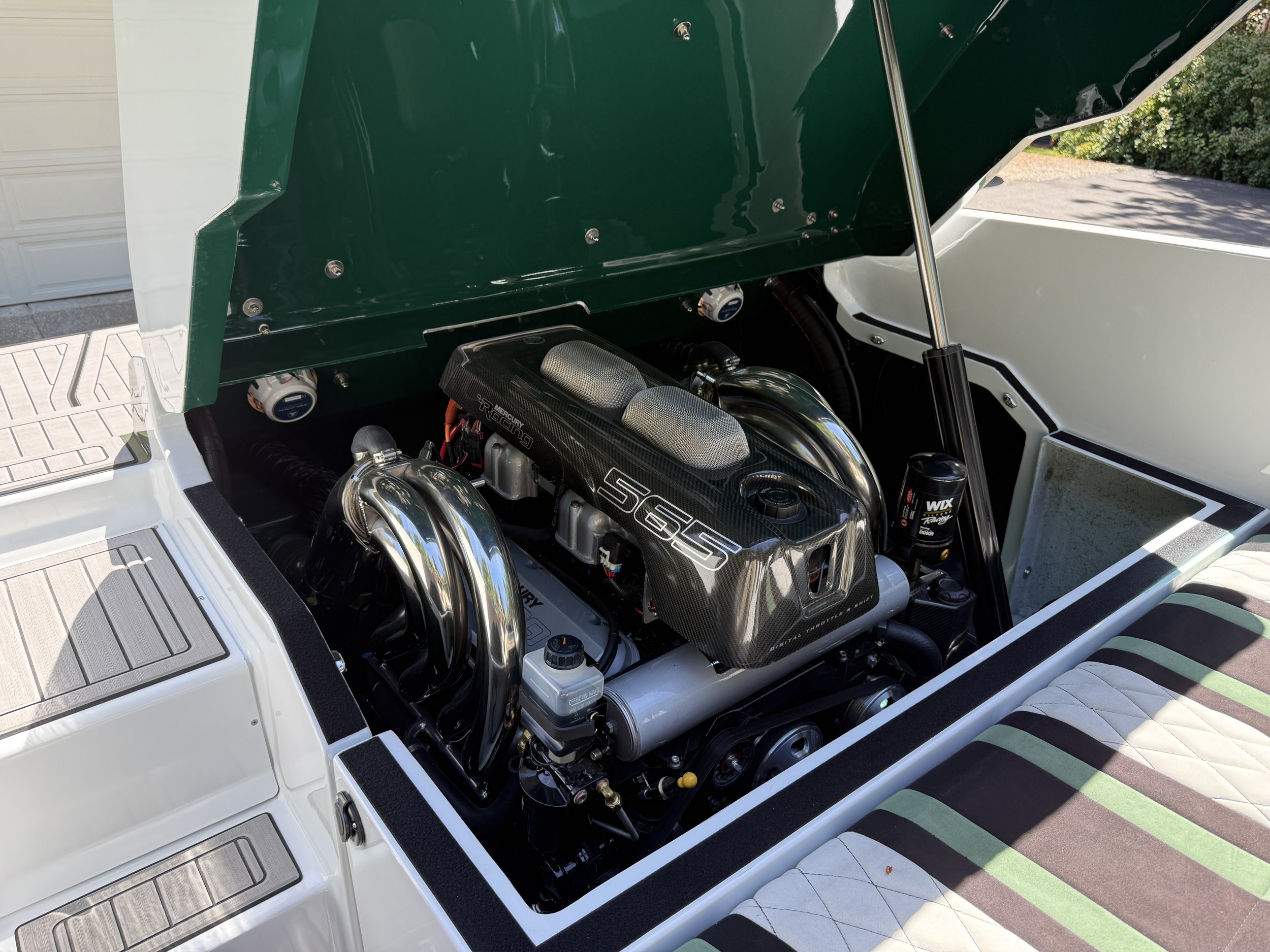 View of a boat engine compartment with a high-performance Mercury Racing engine, silver exhaust headers, and various mechanical components, with part of the boat's deck and seating area visible.