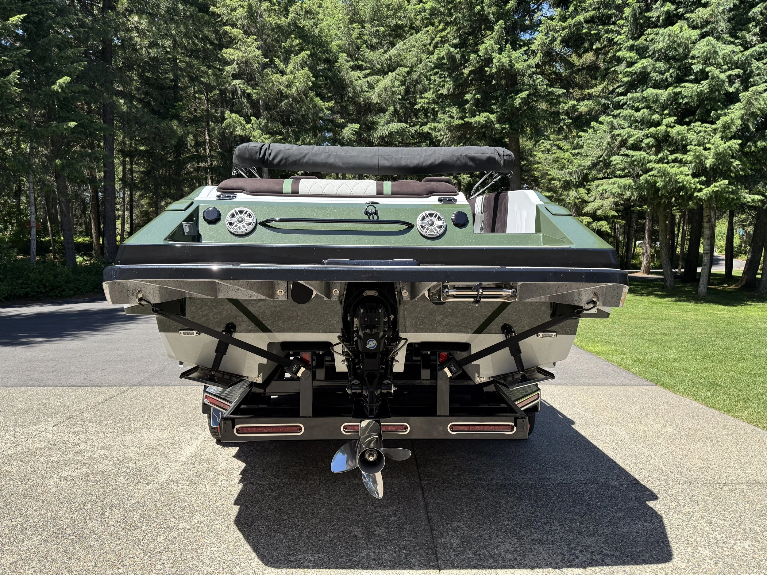 Back view of a boat on a trailer with a motor, parked on a paved surface in front of green trees.