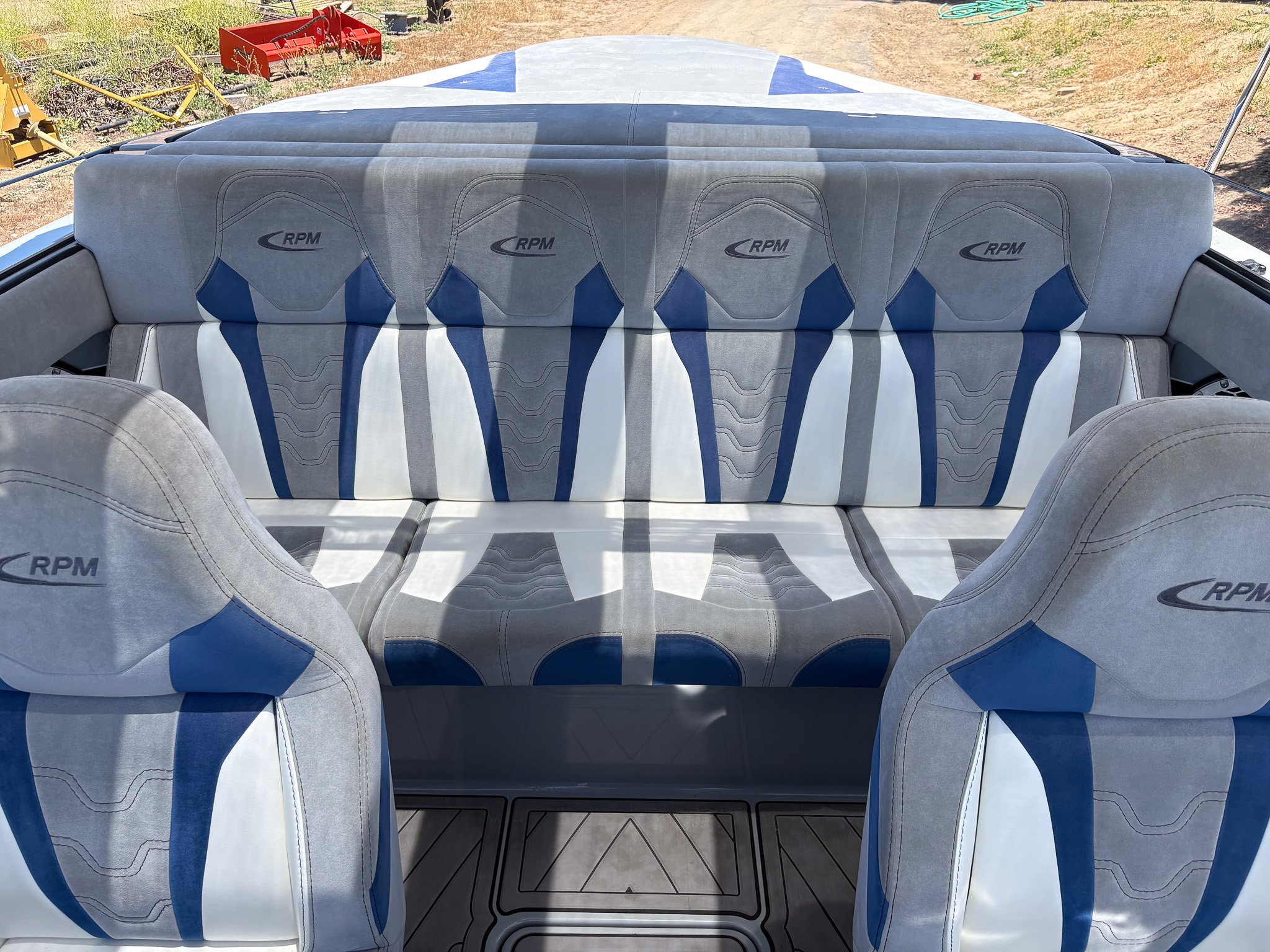 The image shows the back seats of a boat with cushions branded 'RPM', with a white, gray, and blue color scheme, outdoors with a dirt ground and some equipment in the background.