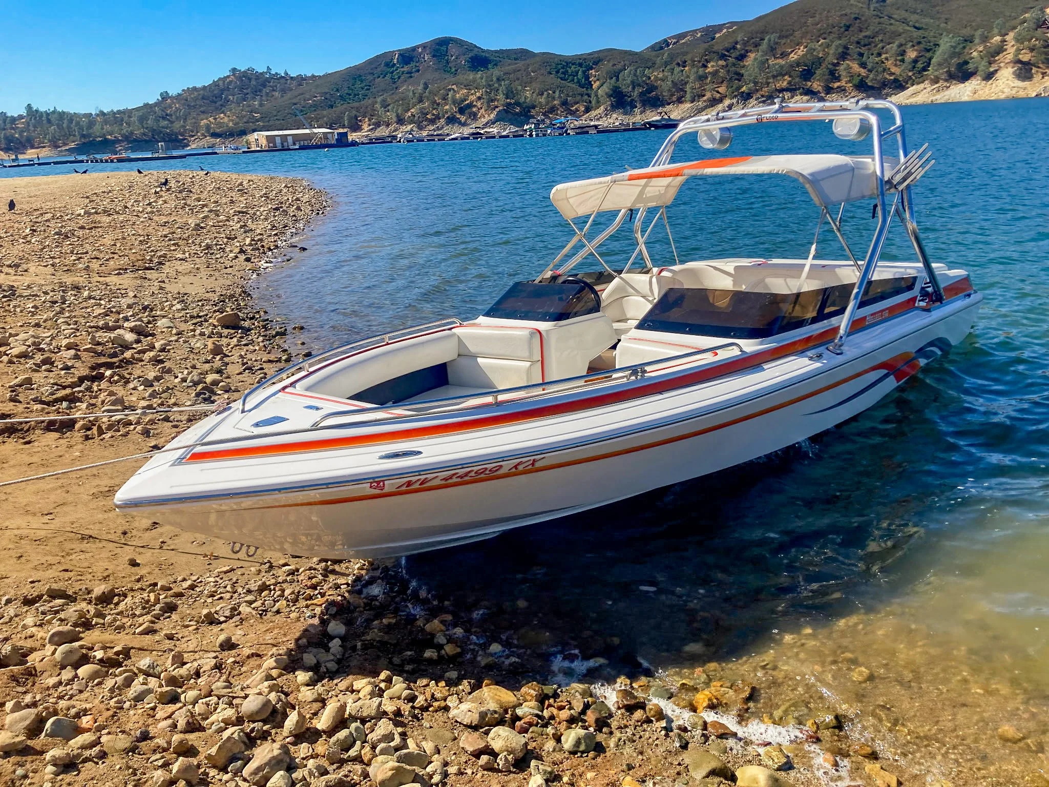 A white speedboat with orange and blue stripes is docked on a rocky shoreline by a lake, with hills and trees in the background under a clear blue sky.