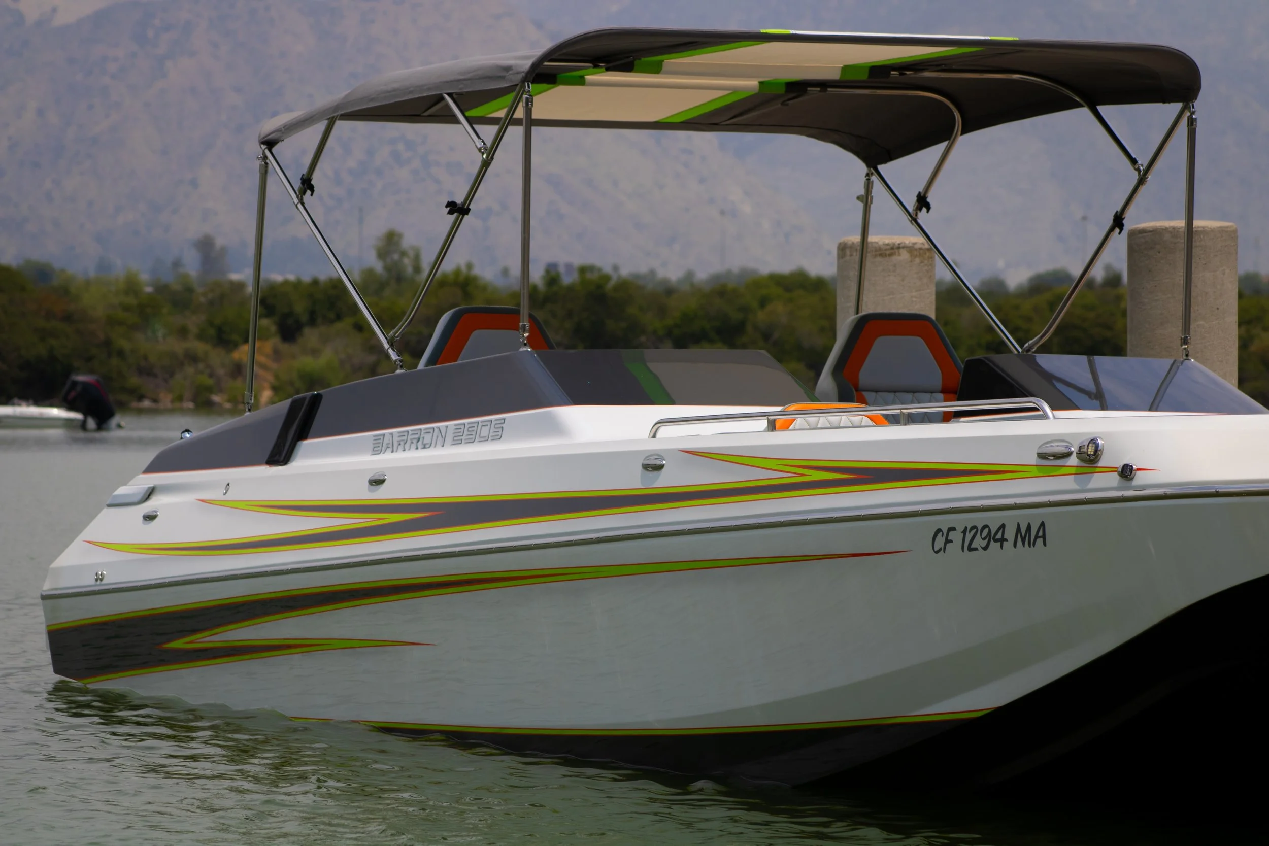 A white and gray speedboat on the water with a black canopy, orange and gray seats, and colorful stripe decals on the side, with trees, mountains, and a small boat in the background.