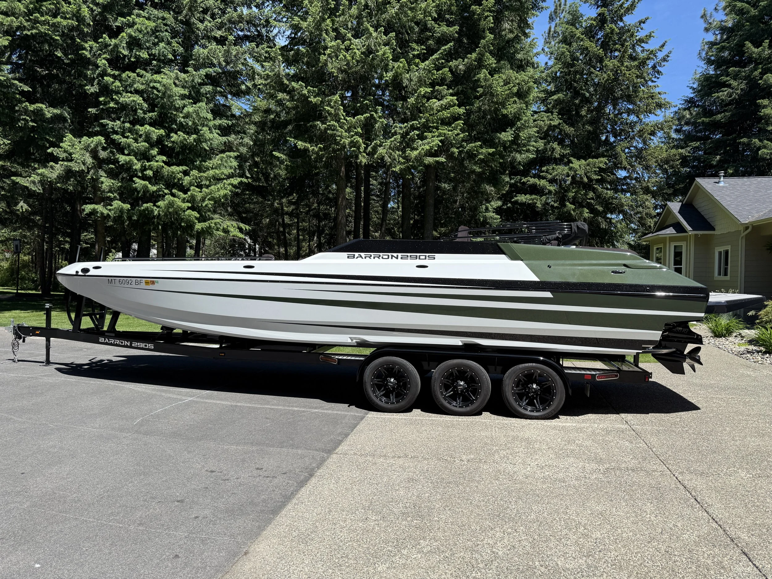 A white boat on a black trailer parked on asphalt, with a house and tall green trees in the background under a clear blue sky.