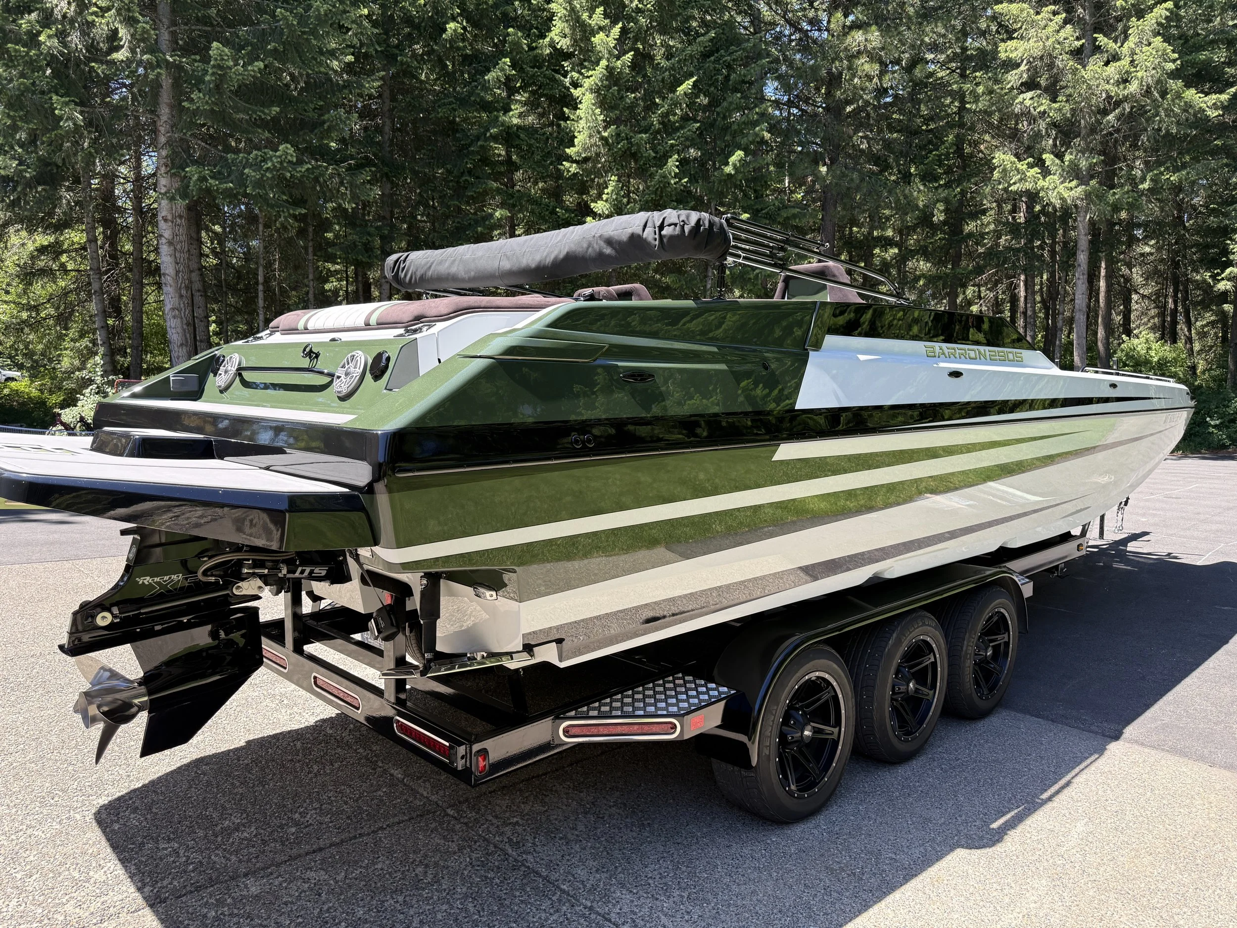 A green and white motorboat on a trailer tailer, with a black covered wakeboard tower, parked on a paved area with a forest of pine trees in the background.