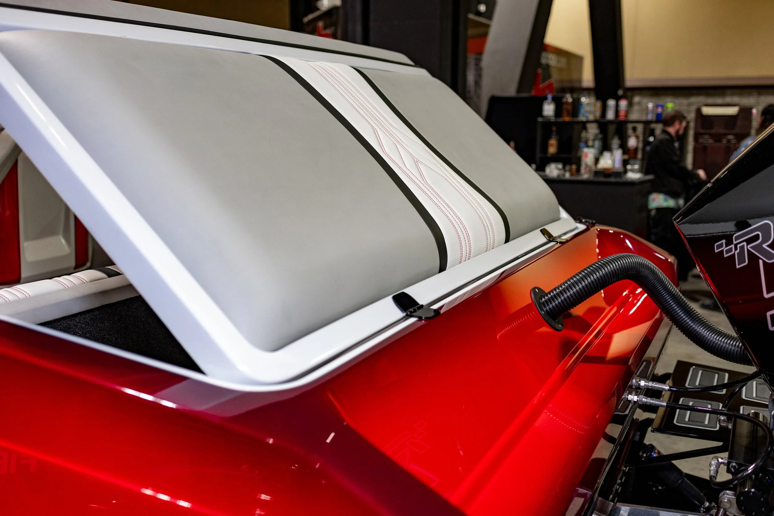 Close-up of a red vehicle with an open compartment revealing a padded, striped cushion and black trim, with a flexible hose connected to some machinery, indoors with a bar and a person in the background.