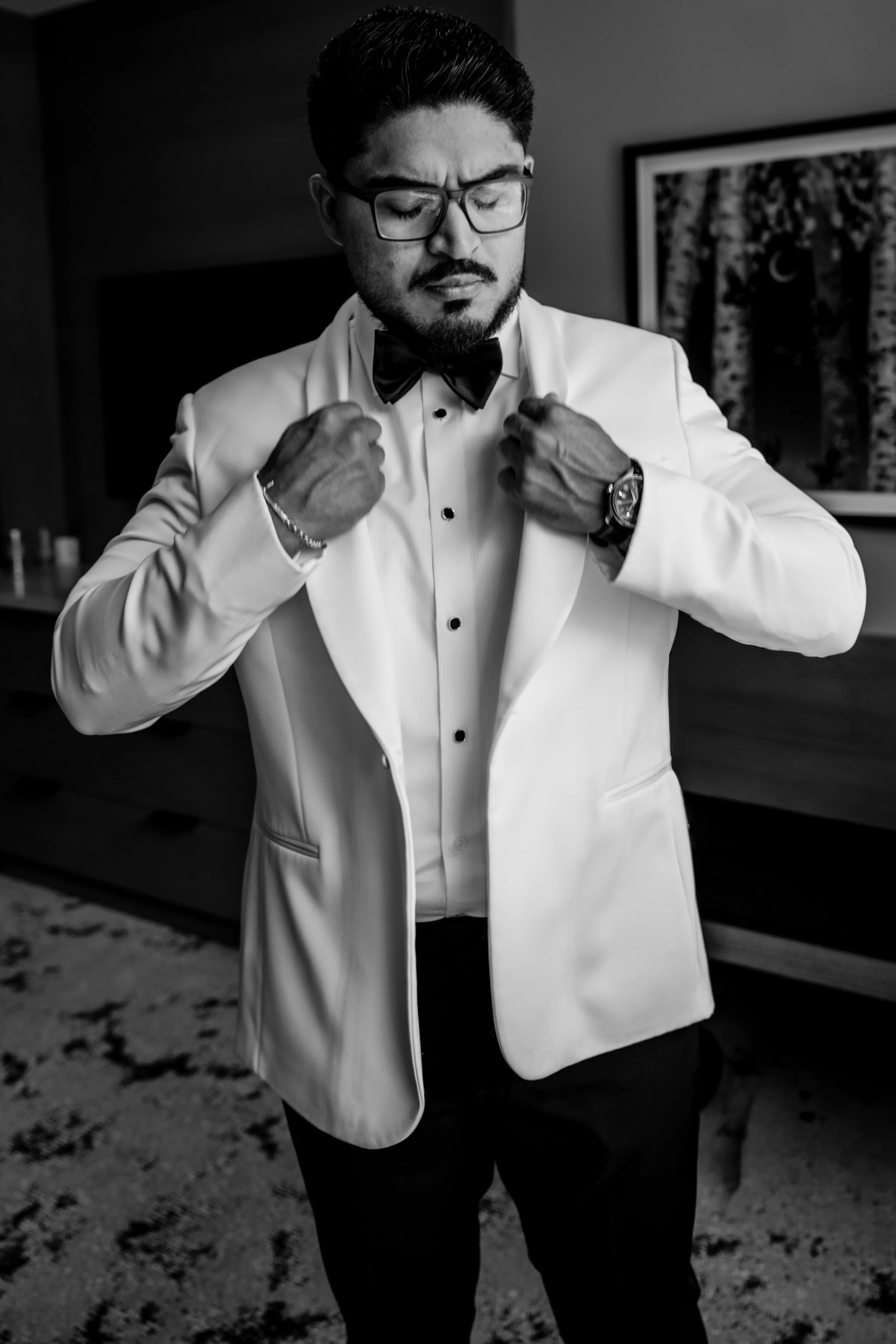 A man in formal attire, wearing a tuxedo with a bow tie, glasses, and a wristwatch, adjusting his jacket collar. He has dark hair, facial hair, and appears to be in a room with a framed picture of trees and a crescent moon on the wall. The image is i