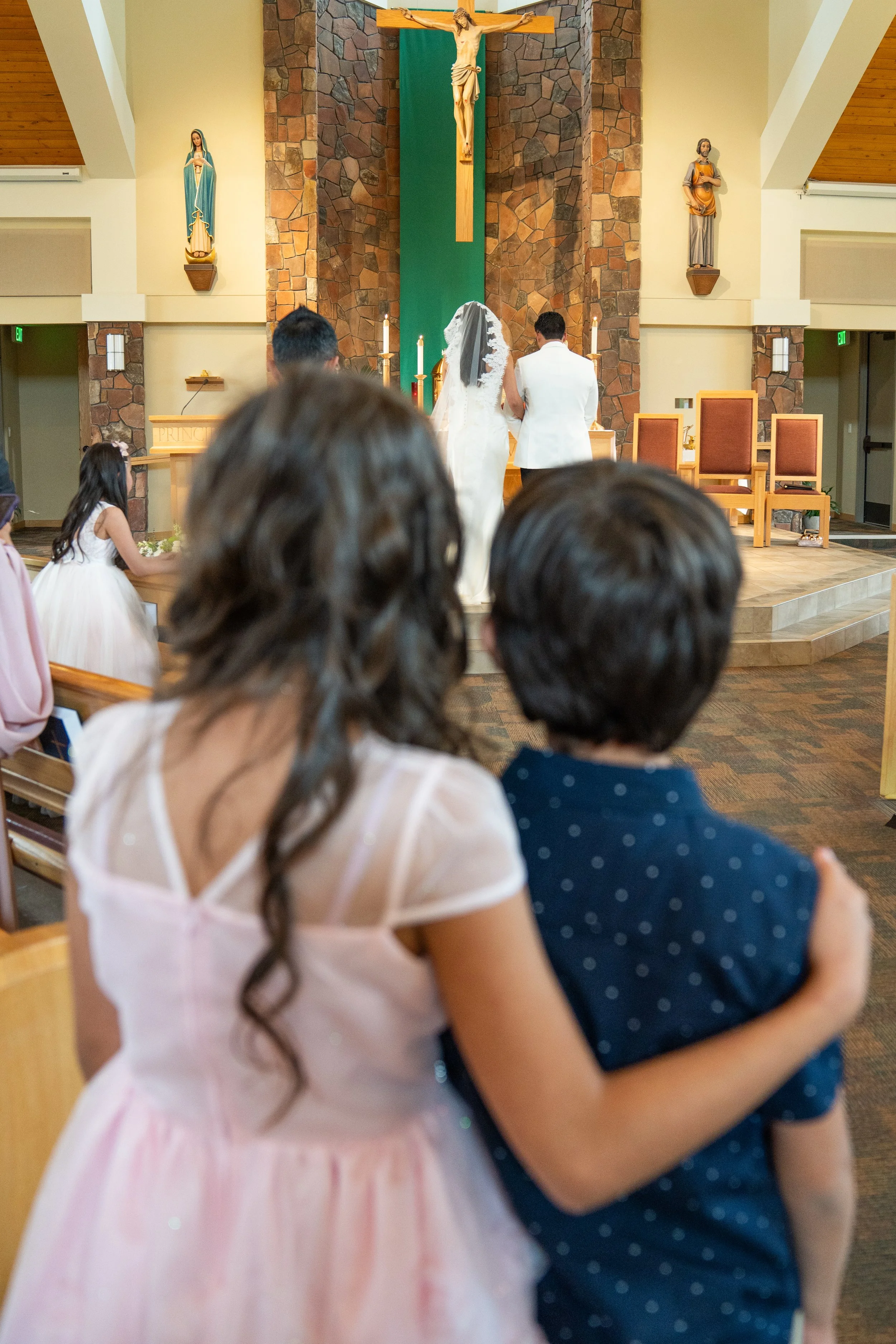 A wedding ceremony taking place in a church with a bride and groom standing at the altar. The altar has candles, and there are religious statues and a crucifix on the wall behind them. Guests are seated and observing the ceremony.