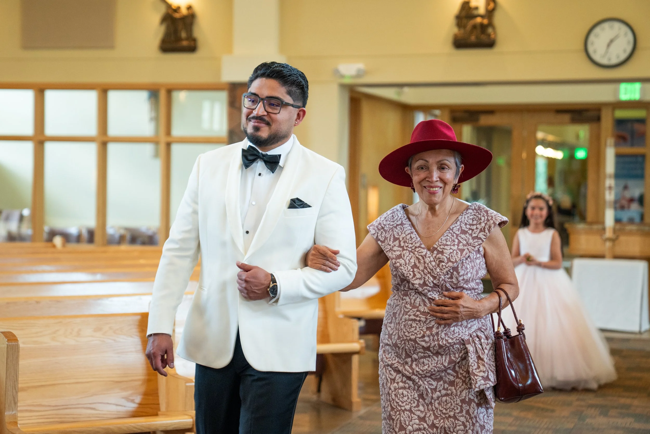 A well-dressed man in a white tuxedo with a black bow tie is walking arm-in-arm with an older woman in a floral dress and large red hat inside a church or event hall. She carries a maroon handbag and is wearing earrings. Behind them, a young girl in 