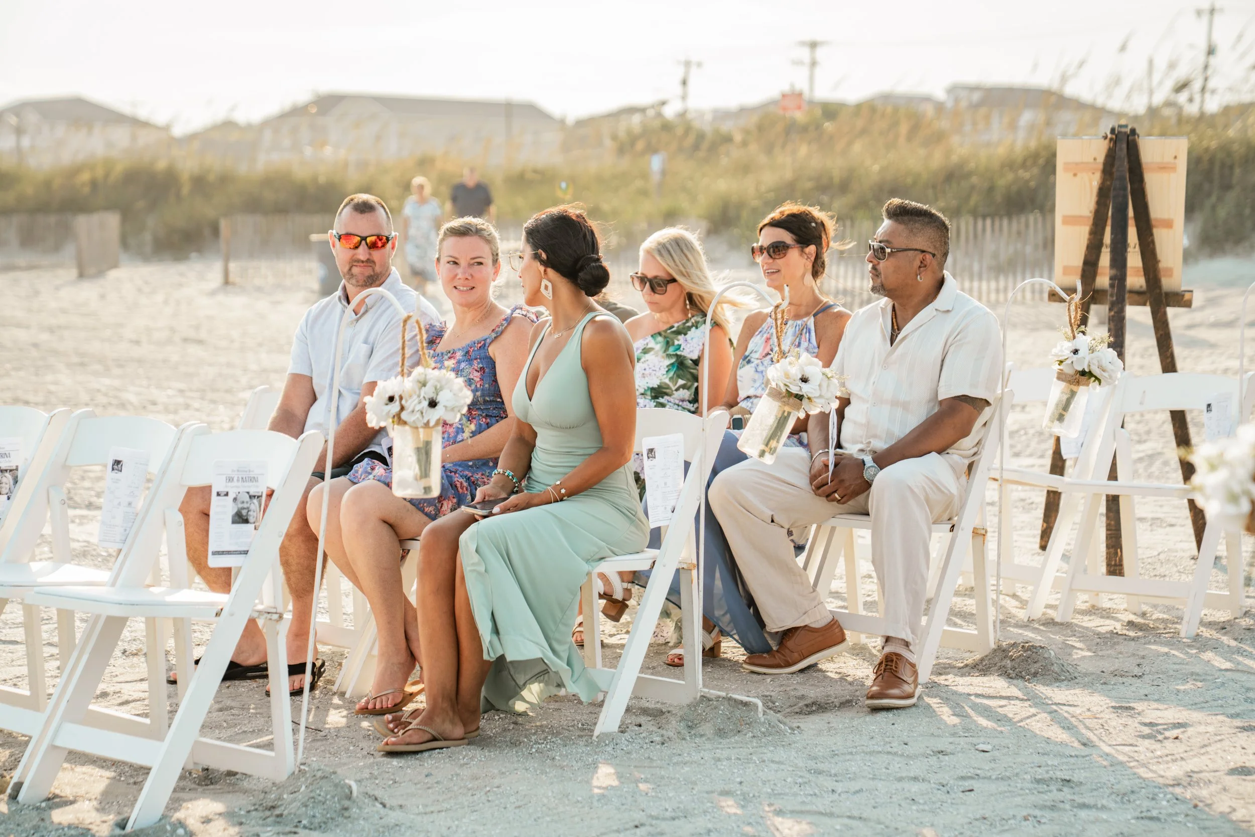Group of diverse people seated on white chairs outdoors, decorated with floral arrangements, at a beach wedding ceremony during daytime.