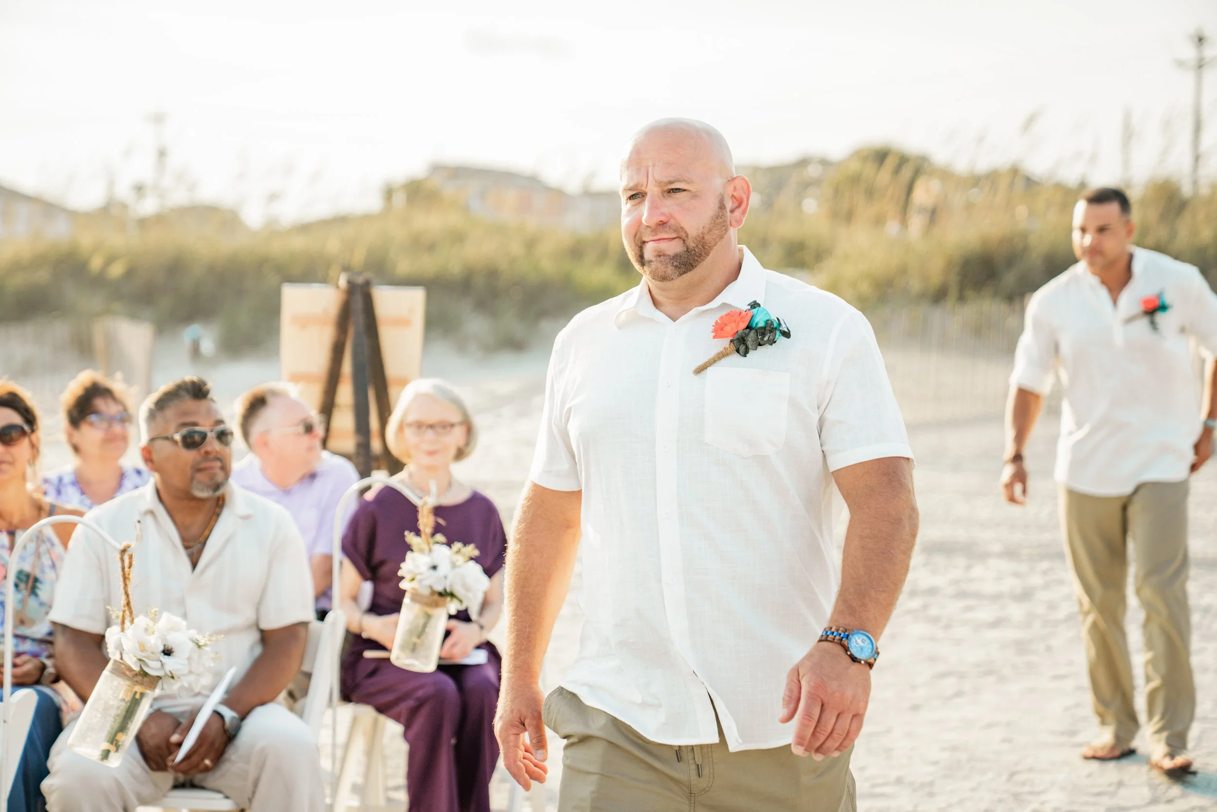 A man with a shaved head and beard wearing a white shirt with a floral boutonniere, walking outdoors during daytime at a beach or park event with guests seated and standing in the background.