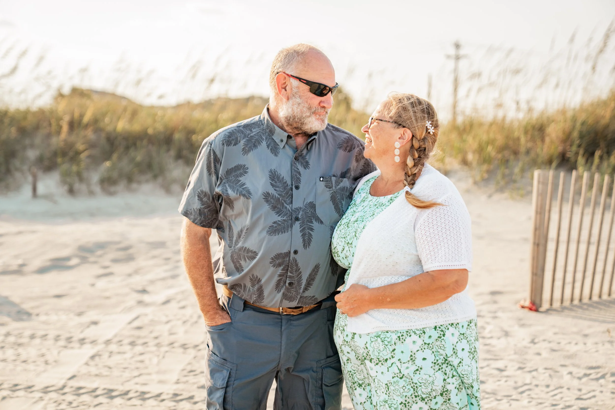 A smiling senior couple standing on a sandy beach during daytime, facing each other and smiling, with dunes and beach grass in the background.