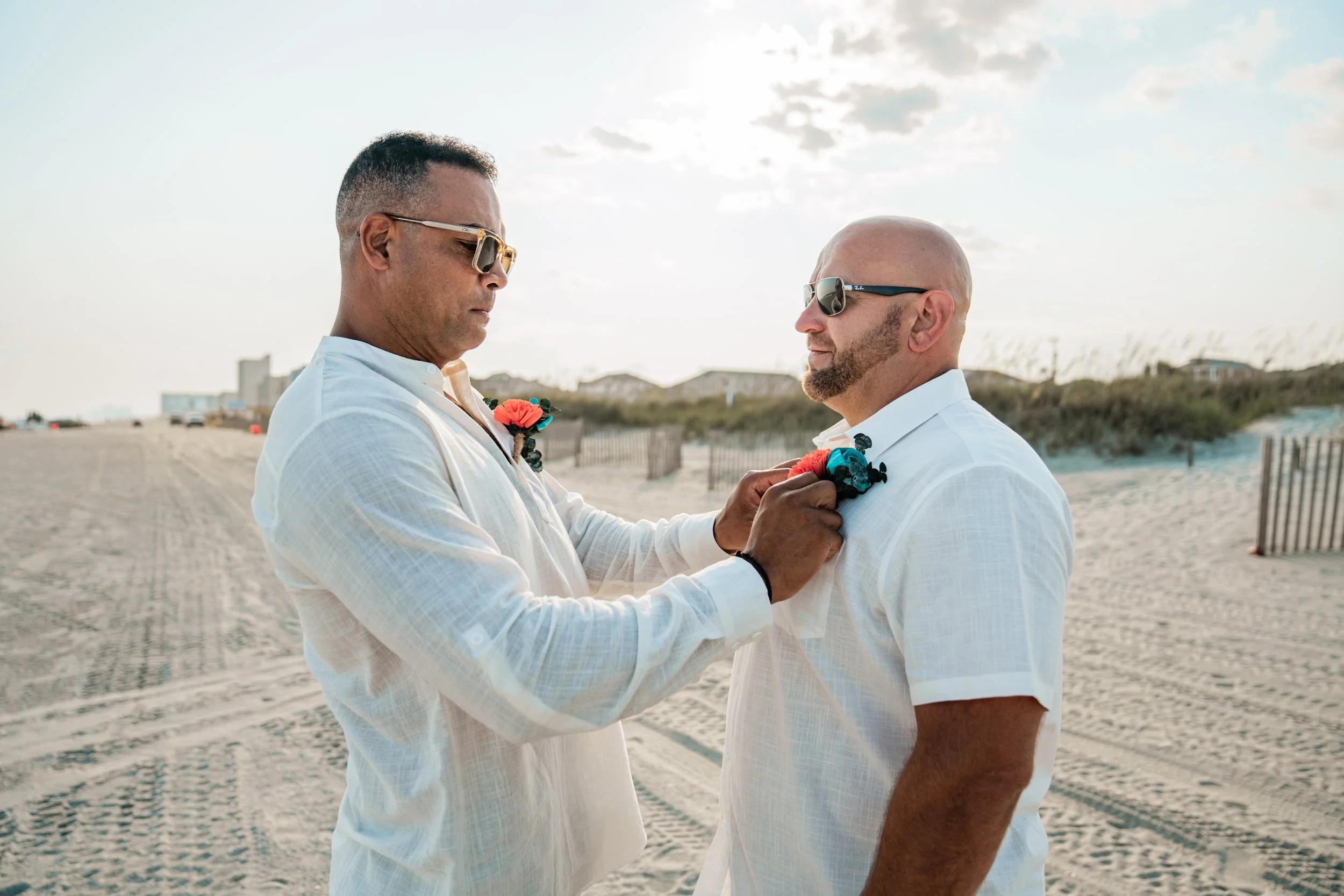 Two men in white shirts exchanging wedding vows on a beach, with a sunset sky and buildings in the background.