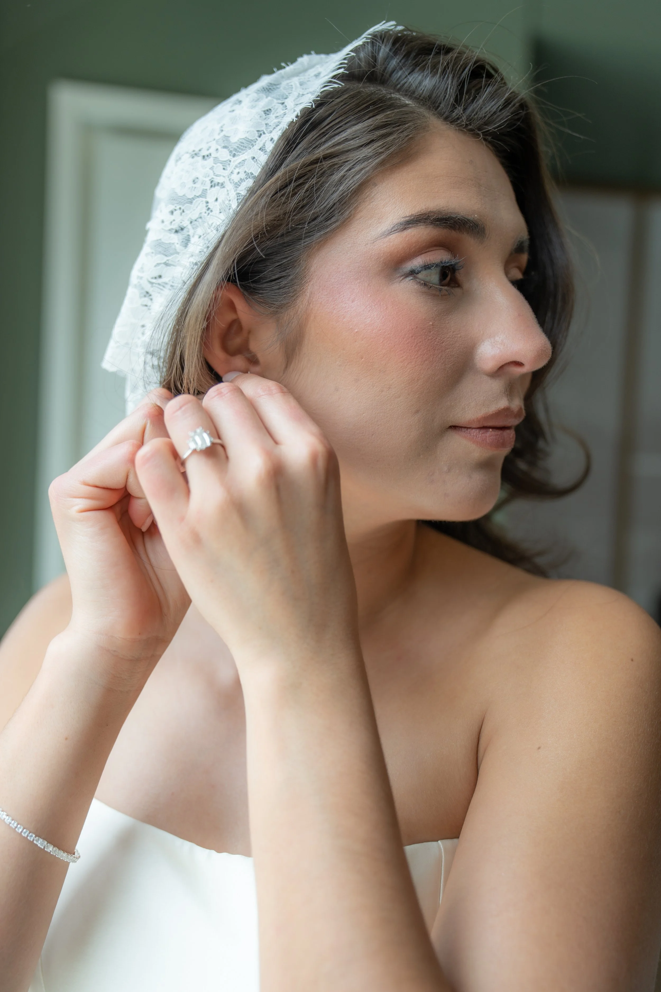 Close-up of a woman with dark hair and light skin, wearing a white lace headscarf, earrings, and a ring, putting on an earring with her right hand.