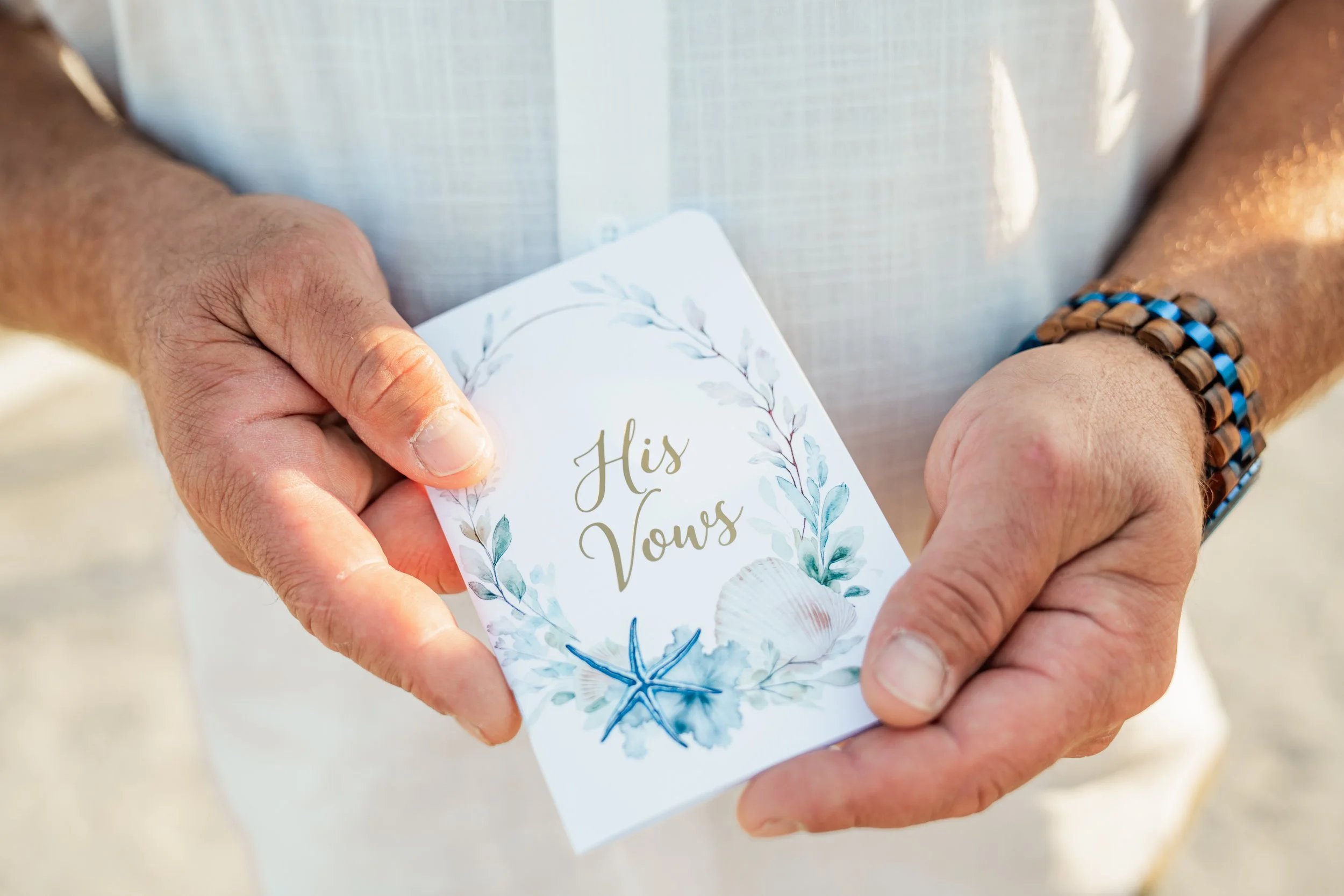Close-up of a person holding a wedding vow card with a floral design, reading 'His Vows'