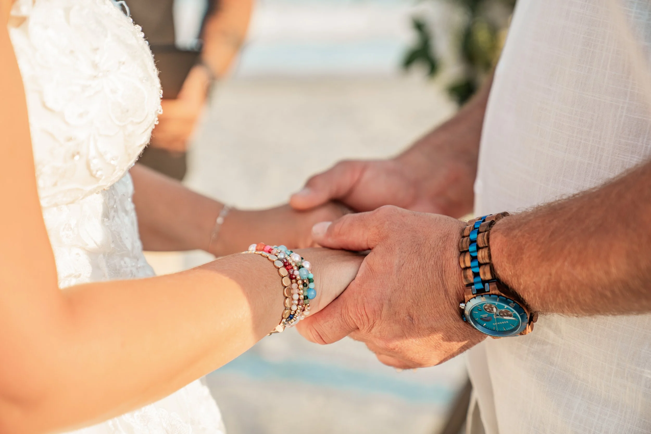 Close-up of a couple holding hands during a wedding ceremony, with focus on their clasped hands and wedding attire. The woman is wearing multiple colorful beaded bracelets, and the man is wearing a watch and a beaded bracelet. The background is blurr