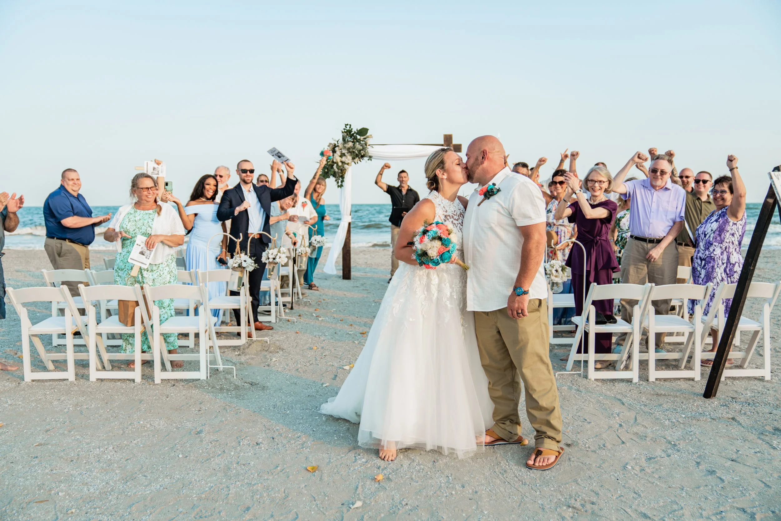 A newlywed couple is sharing a kiss on the beach during their wedding ceremony, with guests cheering and taking photos in the background.