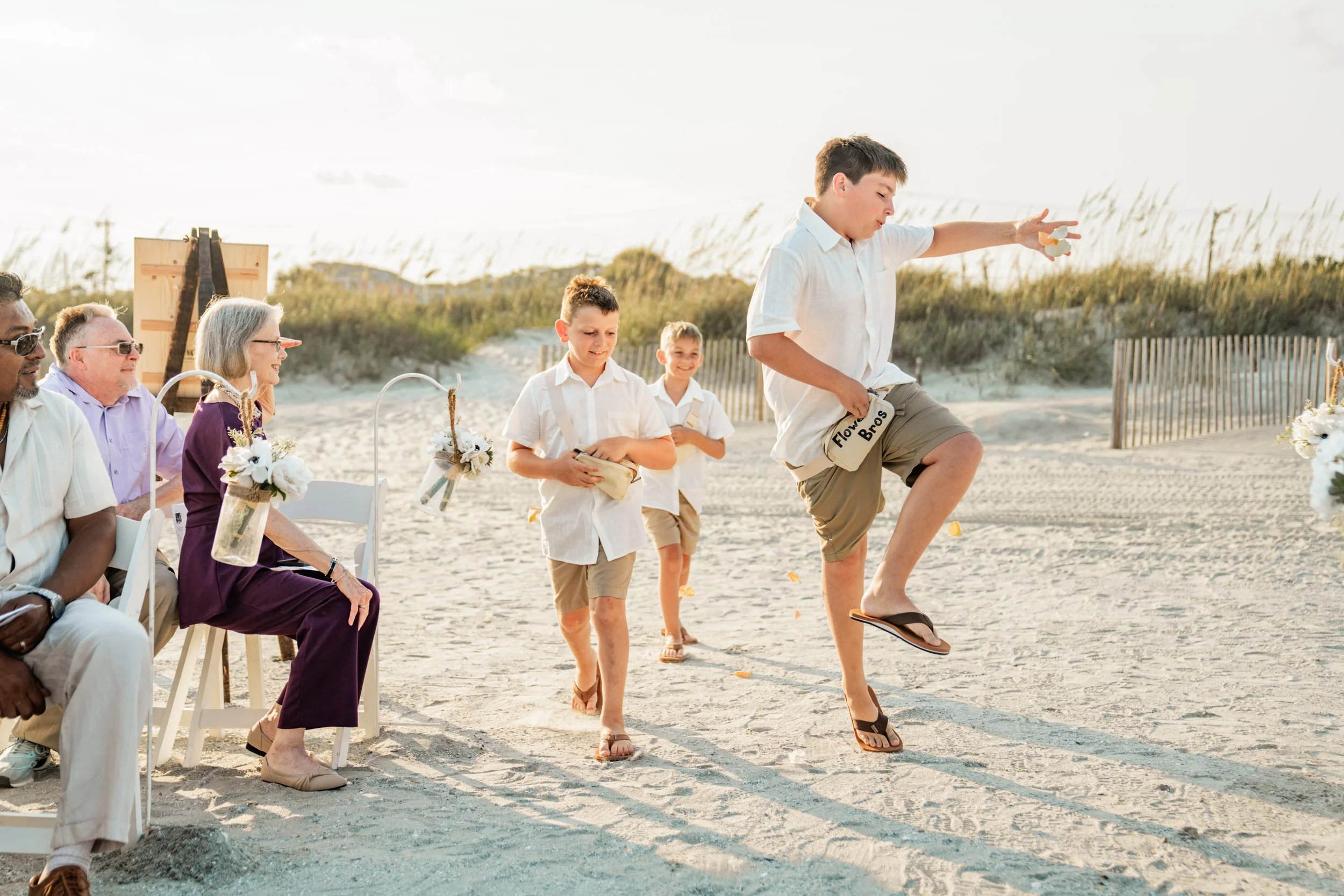 Children in white shirts and khaki shorts participating in a beach wedding ceremony, one boy balancing on a skateboard, adults seated on chairs watching.
