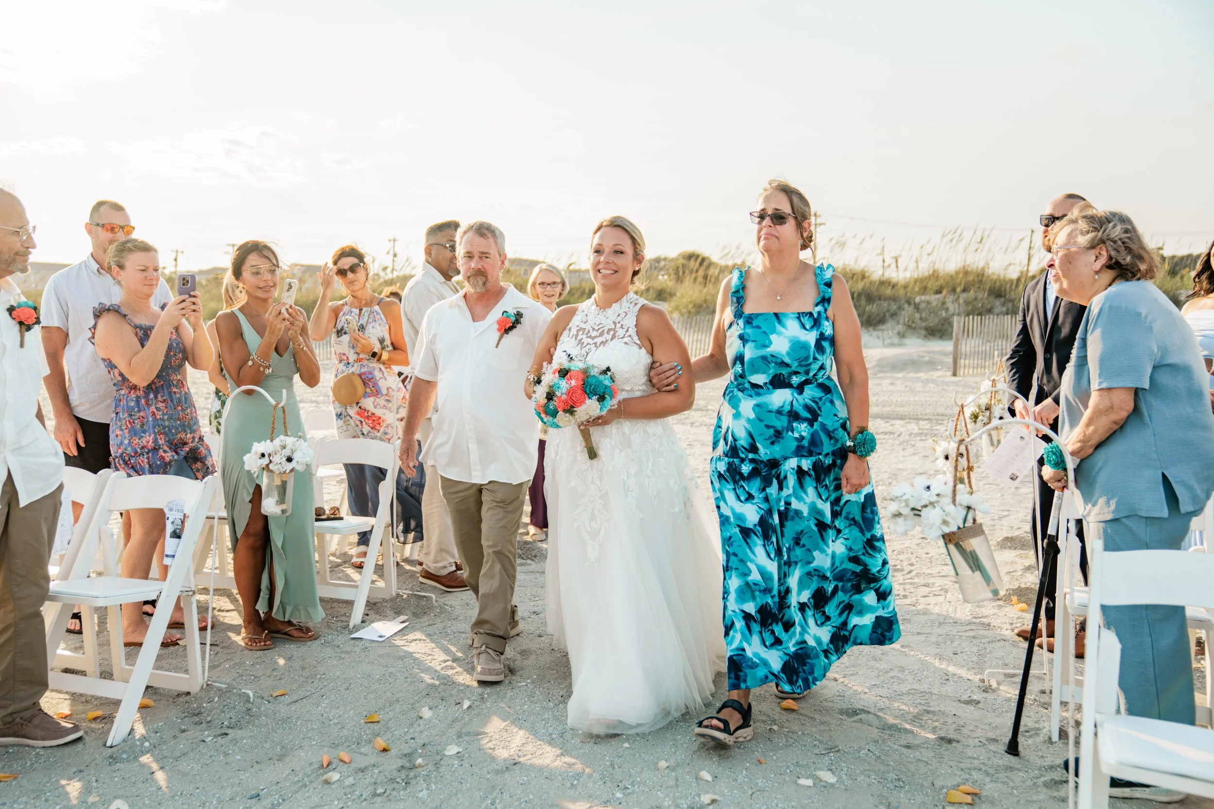 A bride walking down an outdoor beach aisle, accompanied by a woman in a blue dress, with guests standing and sitting around, some taking photos, in daylight conditions.