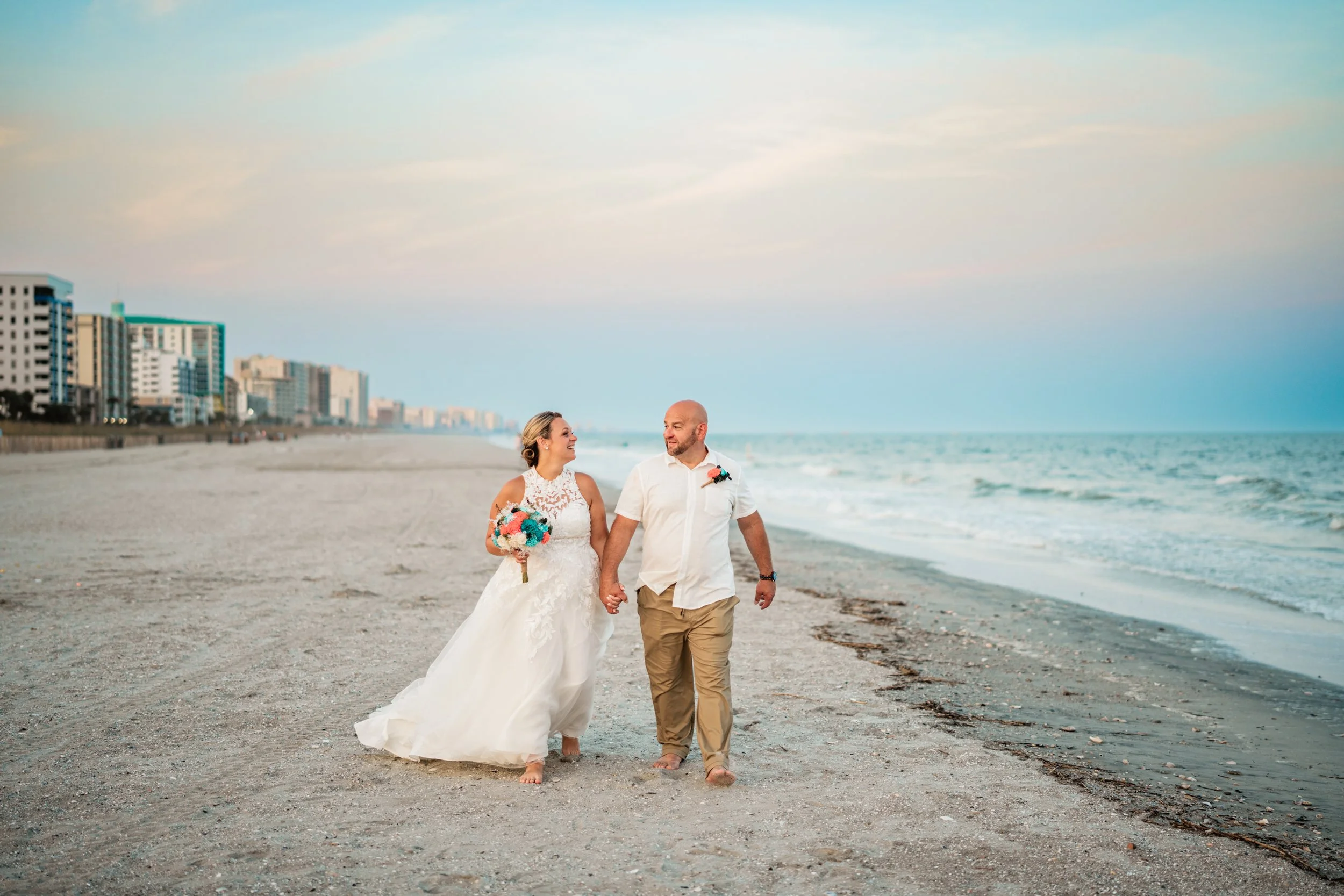 A newlywed couple walking barefoot on the beach, holding hands. The bride is in a white lace wedding dress holding a colorful bouquet and looking at the groom. The groom is in a light-colored shirt and khaki pants, looking at the bride. They are smil