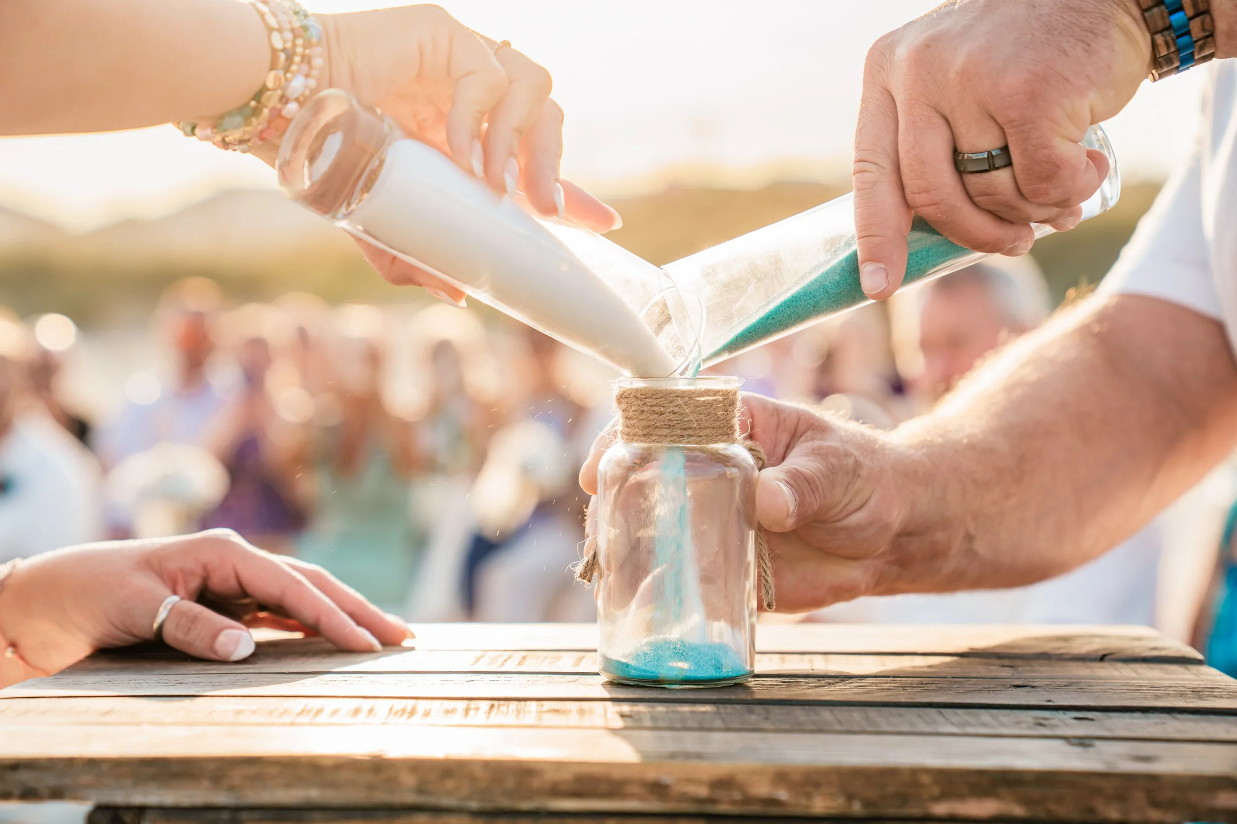 People pouring white and blue sand into a jar, participating in a sand ceremony outdoors during sunset.