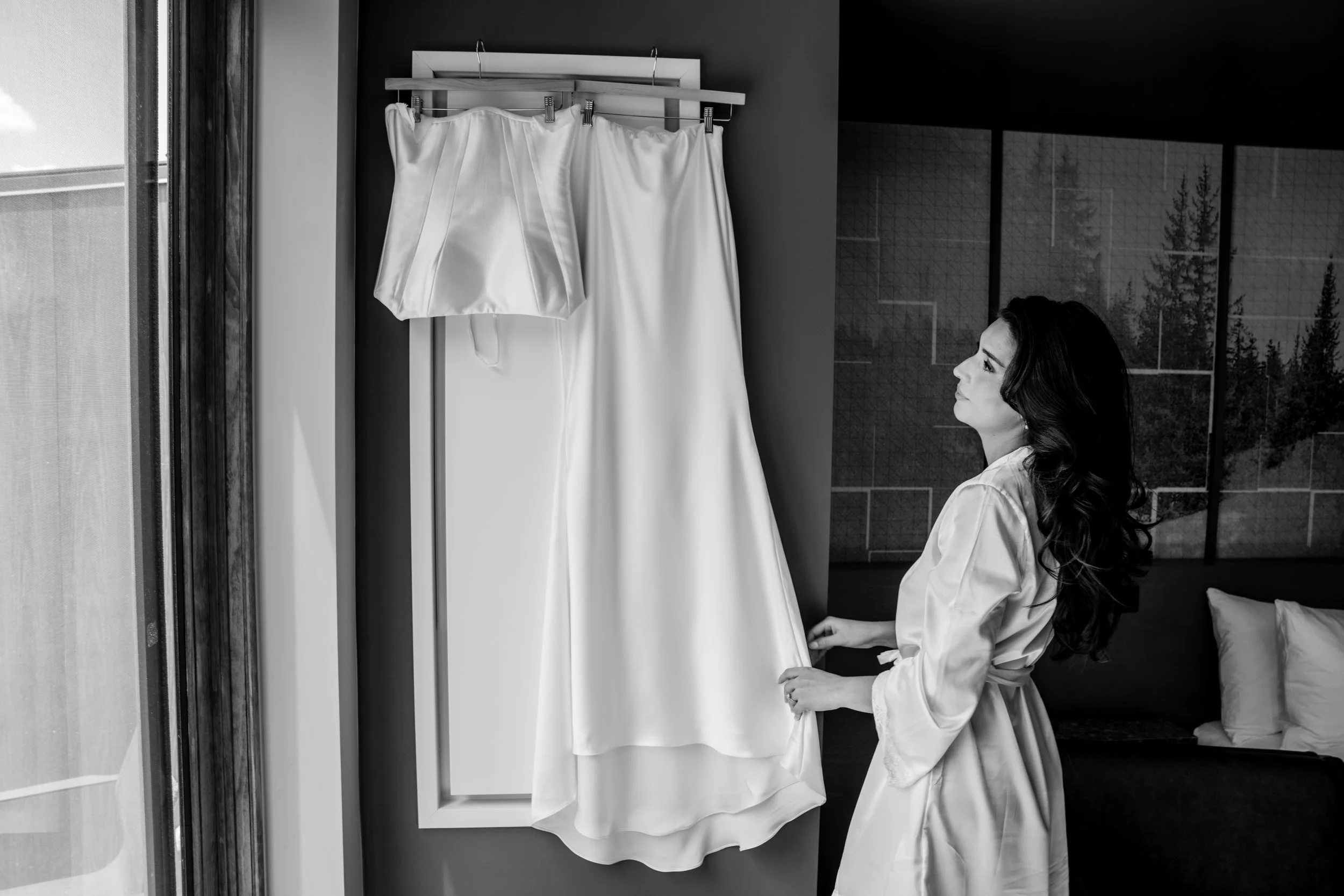 A woman with long dark hair, dressed in a white silk robe, is sitting on a bed while looking at a white wedding dress and a matching silk robe hanging on a hanger in front of her.