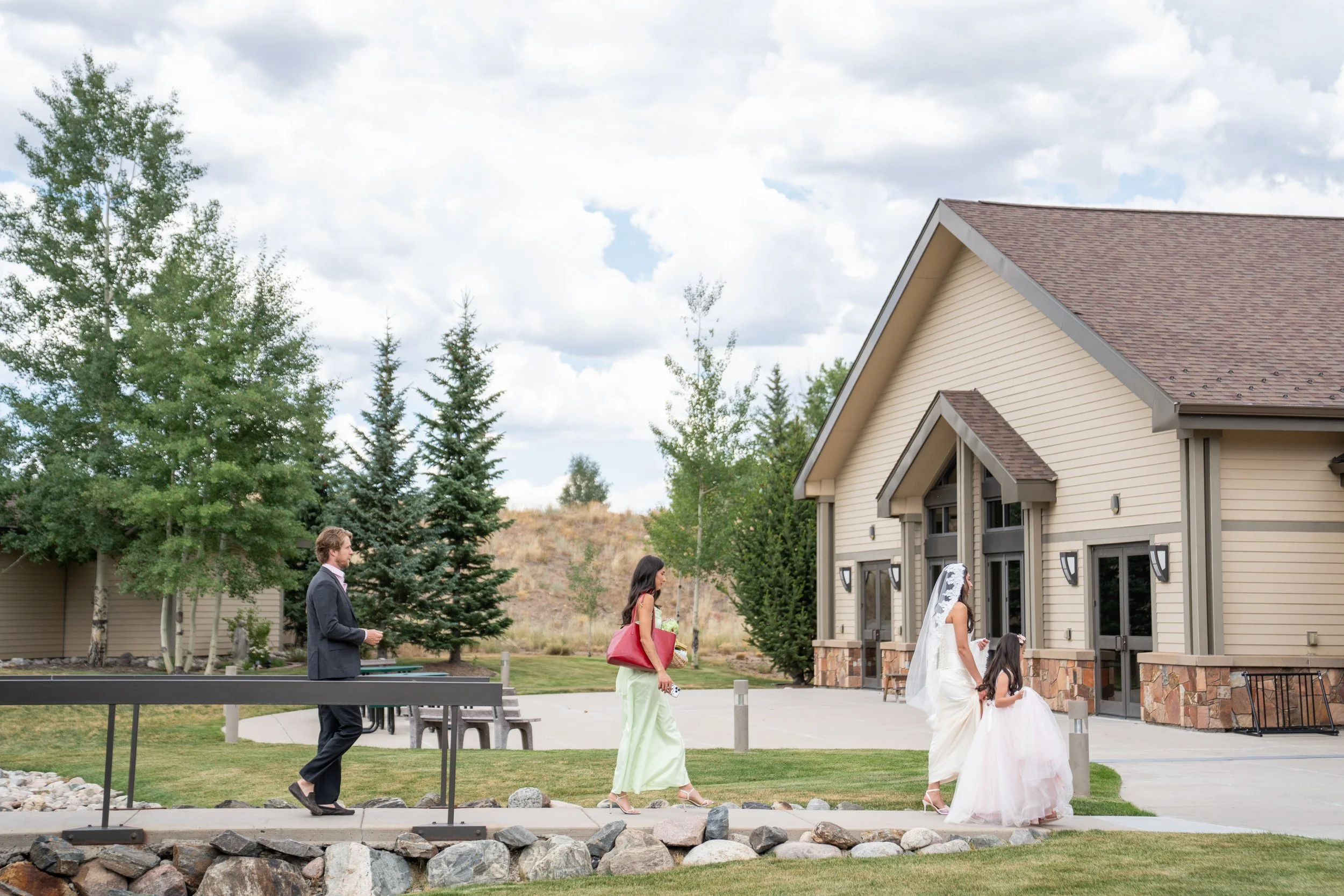 People attending a wedding ceremony outdoors near a beige house with a brown roof, with a backdrop of trees and cloudy sky.