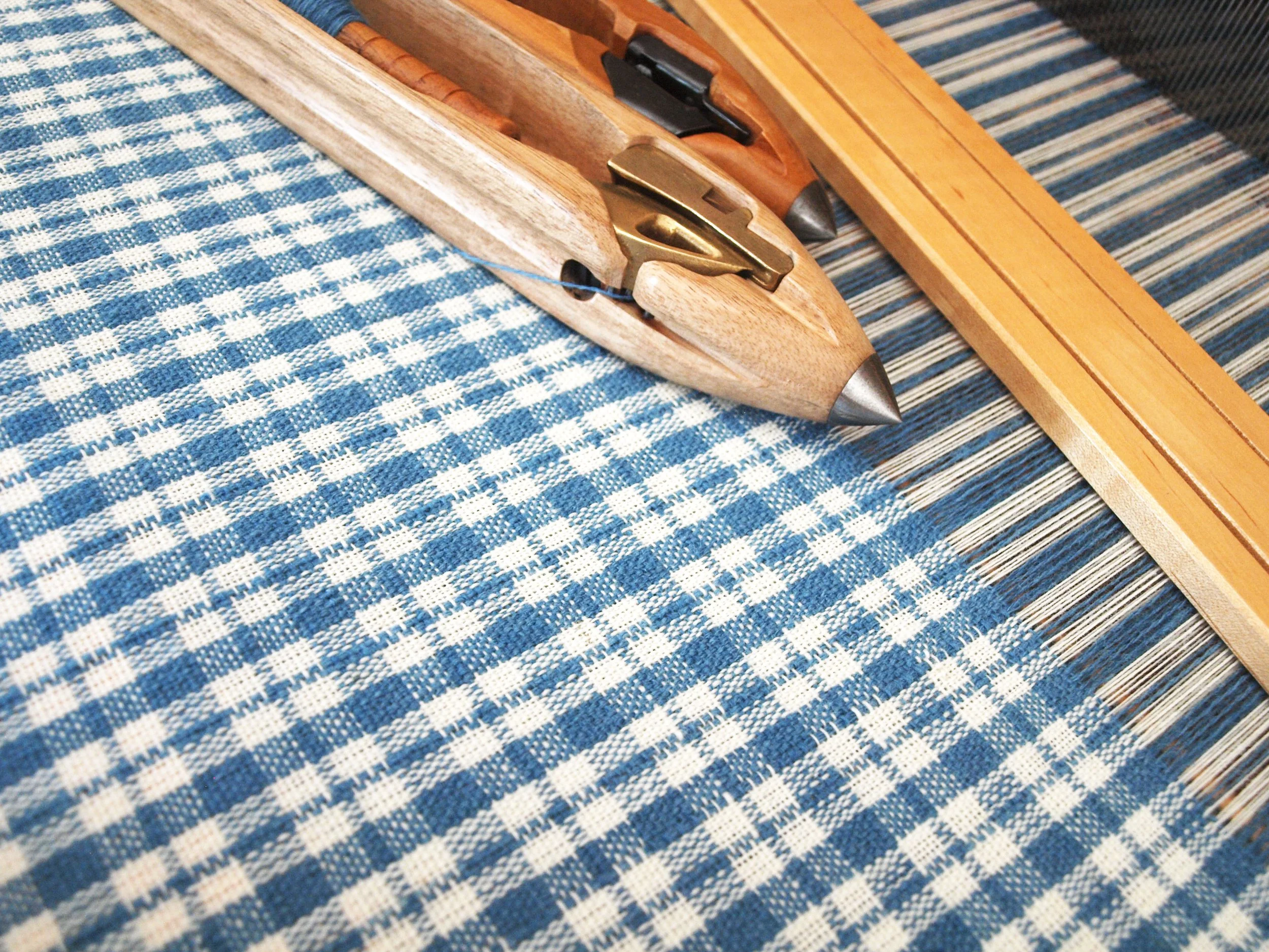 A light blue indigo and creamy white wool flannel check-patterned fabric on the loom with two shuttles resting on top.
