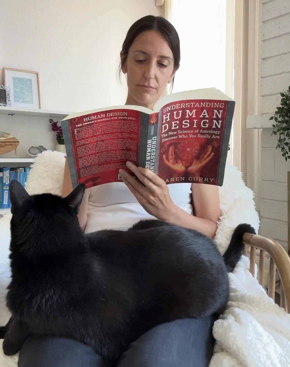 Woman reading "Understanding Human Design" by Karen Curry while a black cat rests on her lap