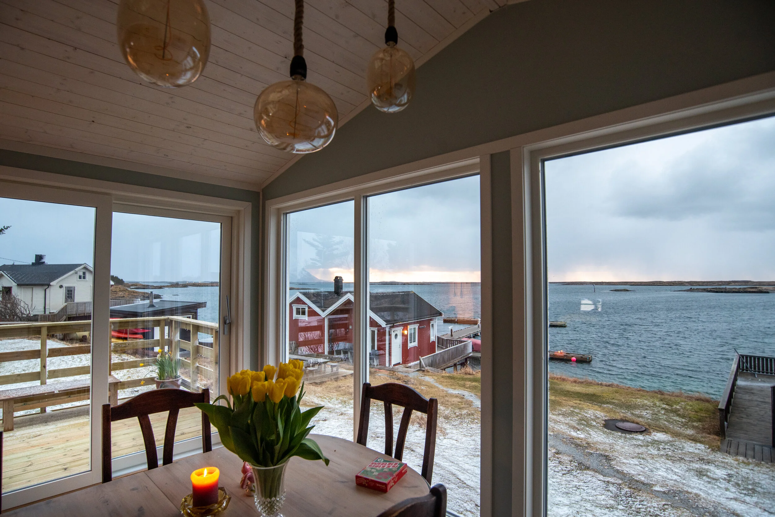 Innendørs bilde av et spisebord med blomster og et lys, med store vinduer som viser en utsikt over sjøen og hus utendørs.