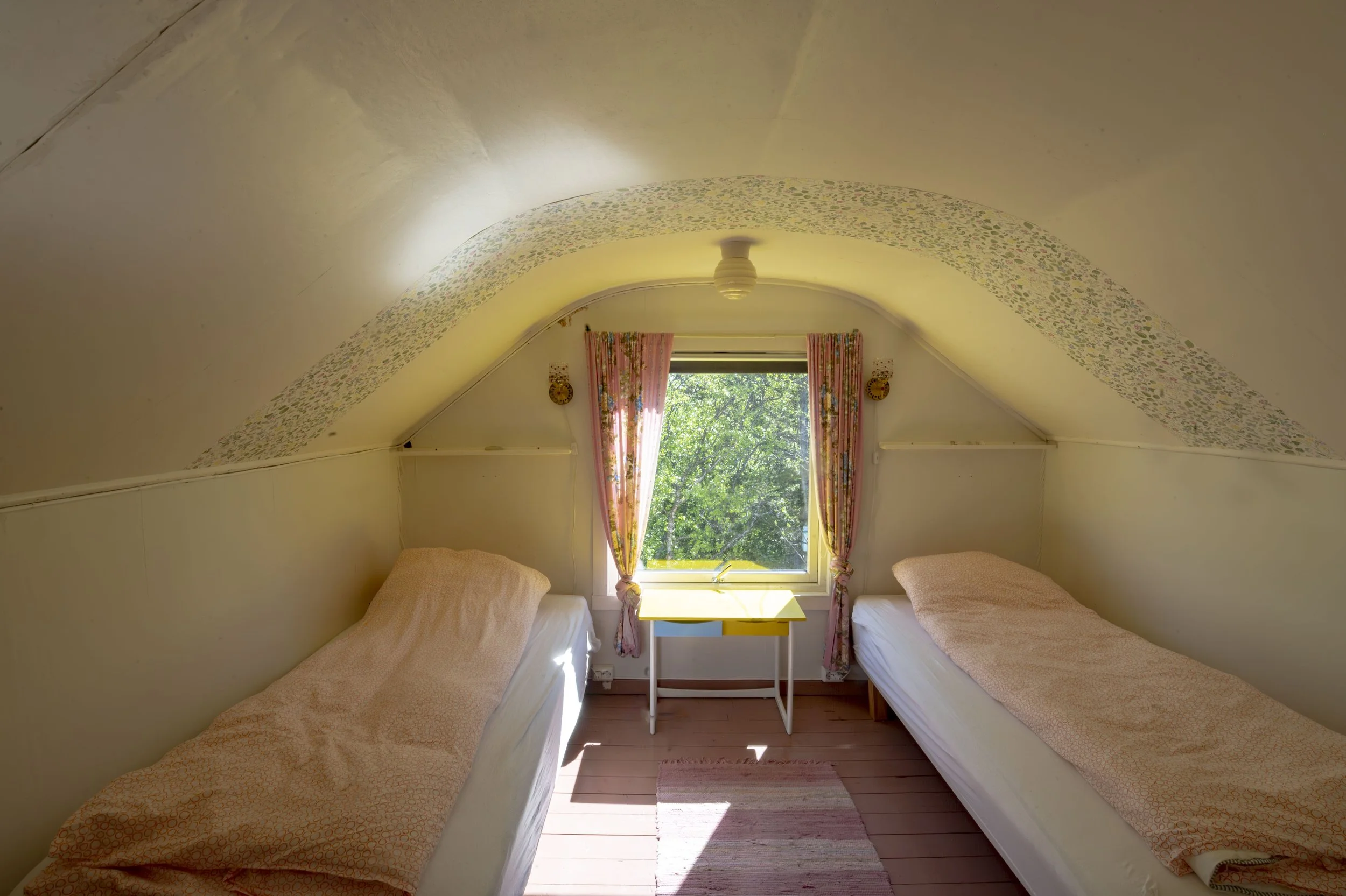 To enkeltsenger med beige sengetøy i et rom med skråtak. Det er et lite vindu med blomstrede gardiner og utsikt til grønne trær. Midt i bildet står et lite hvitt bord.
