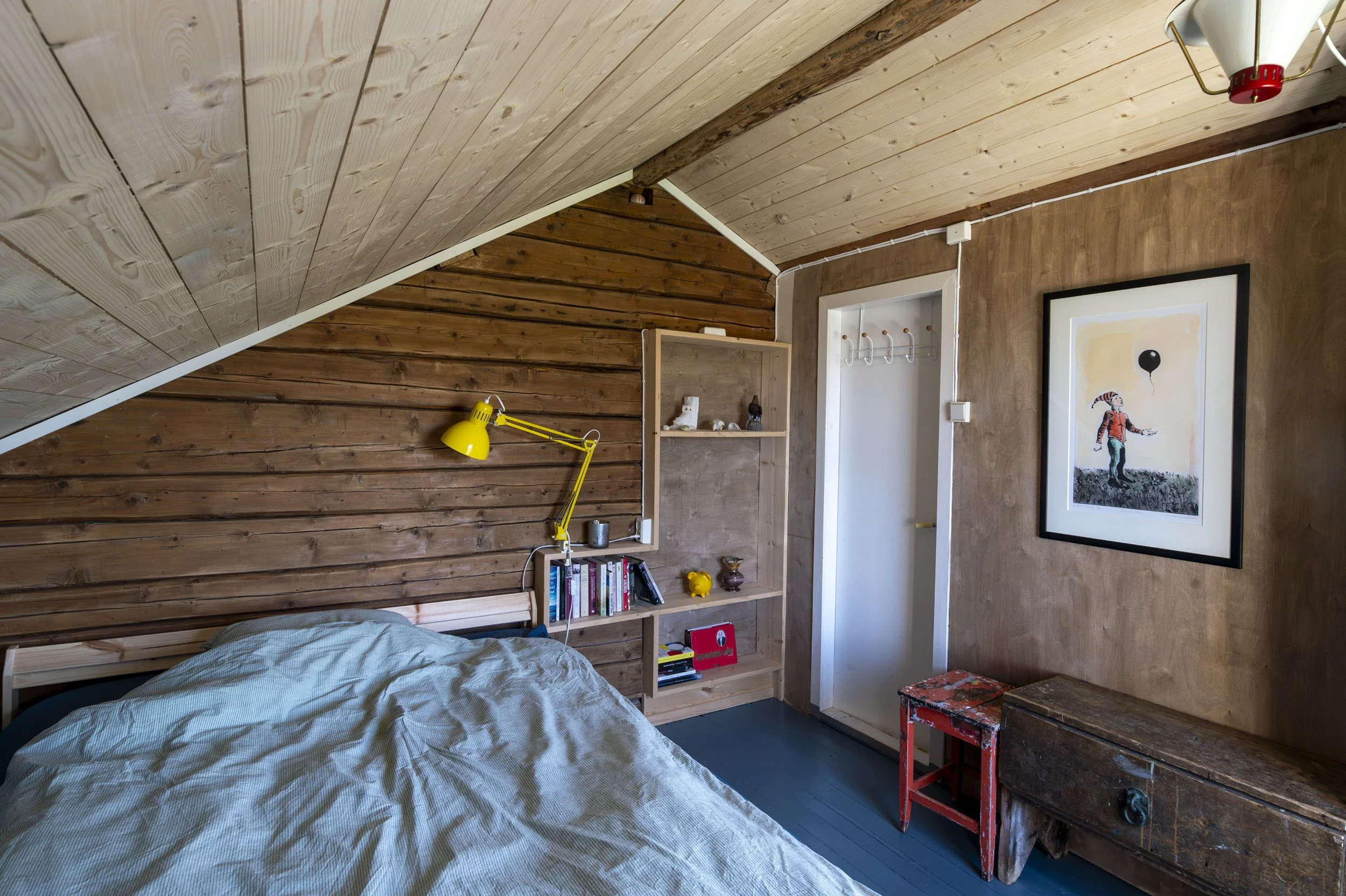 Et soverom med skrå tak, trevegger, seng med lyseblå sengetak, bokhylle med bøker og dekorasjoner, et bilde av en person med ballong, og et møbel laget av gamle treplanker.