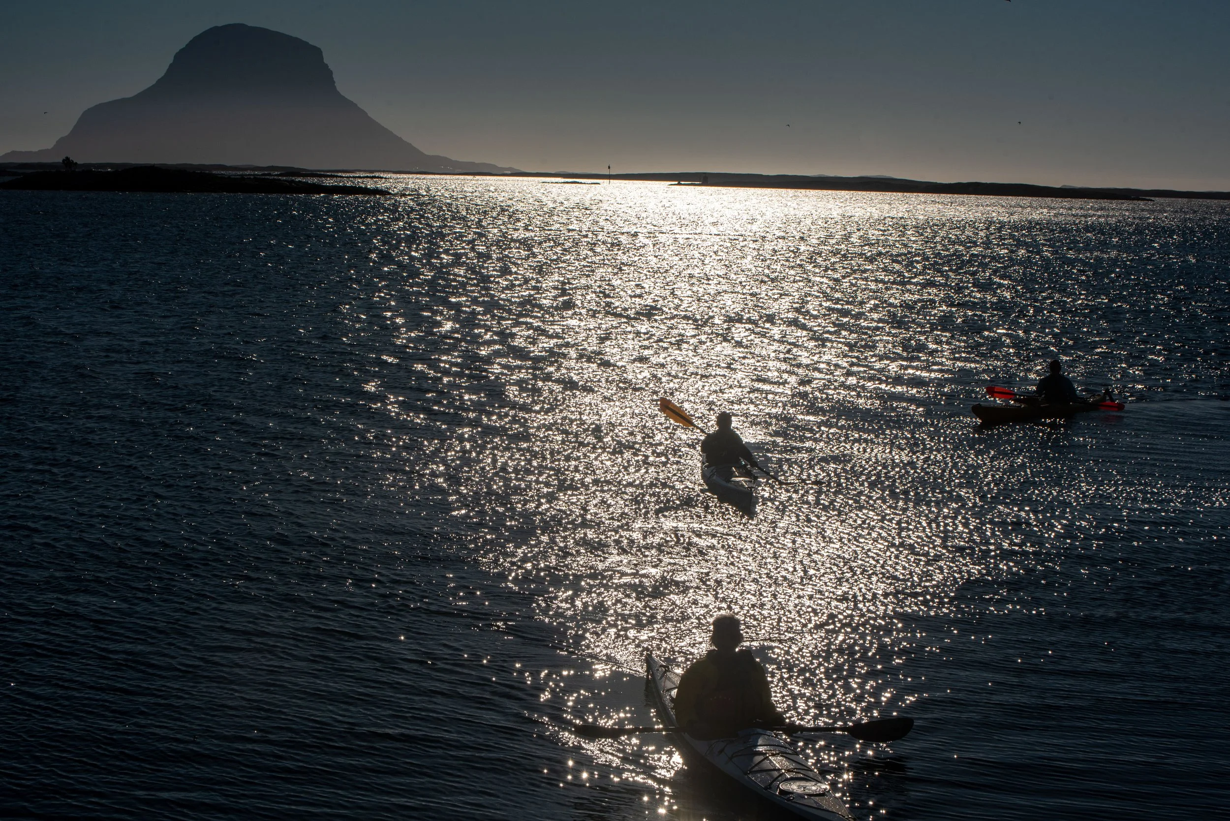 To personer som padler kajakk i sjøen med fjell i bakgrunnen under solnedgang, og lysrefleksjoner på vannet.