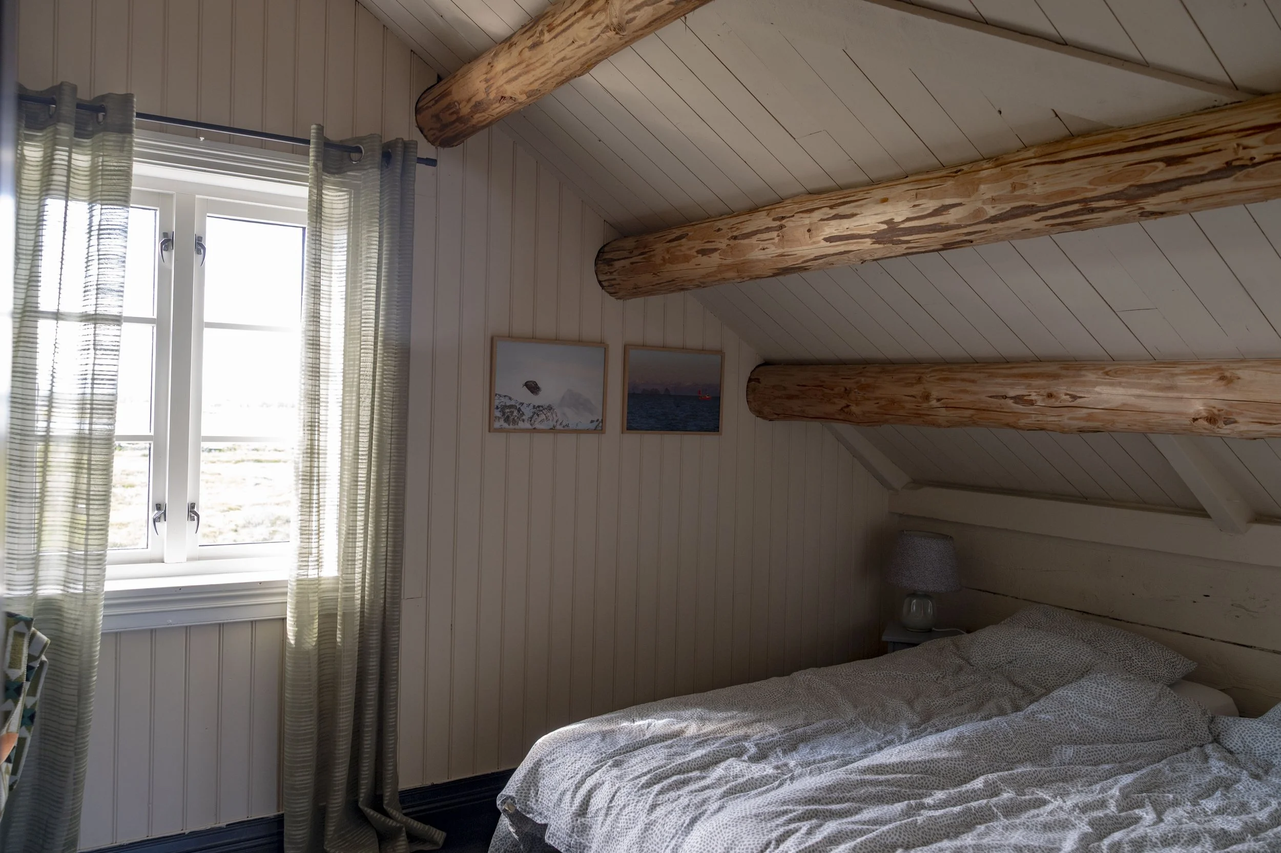 Et soverom med to store, runde trebjelker på taket, hvite panelvegger og et vindu med grønne og hvite gardiner. Det henger to bilder på veggen.