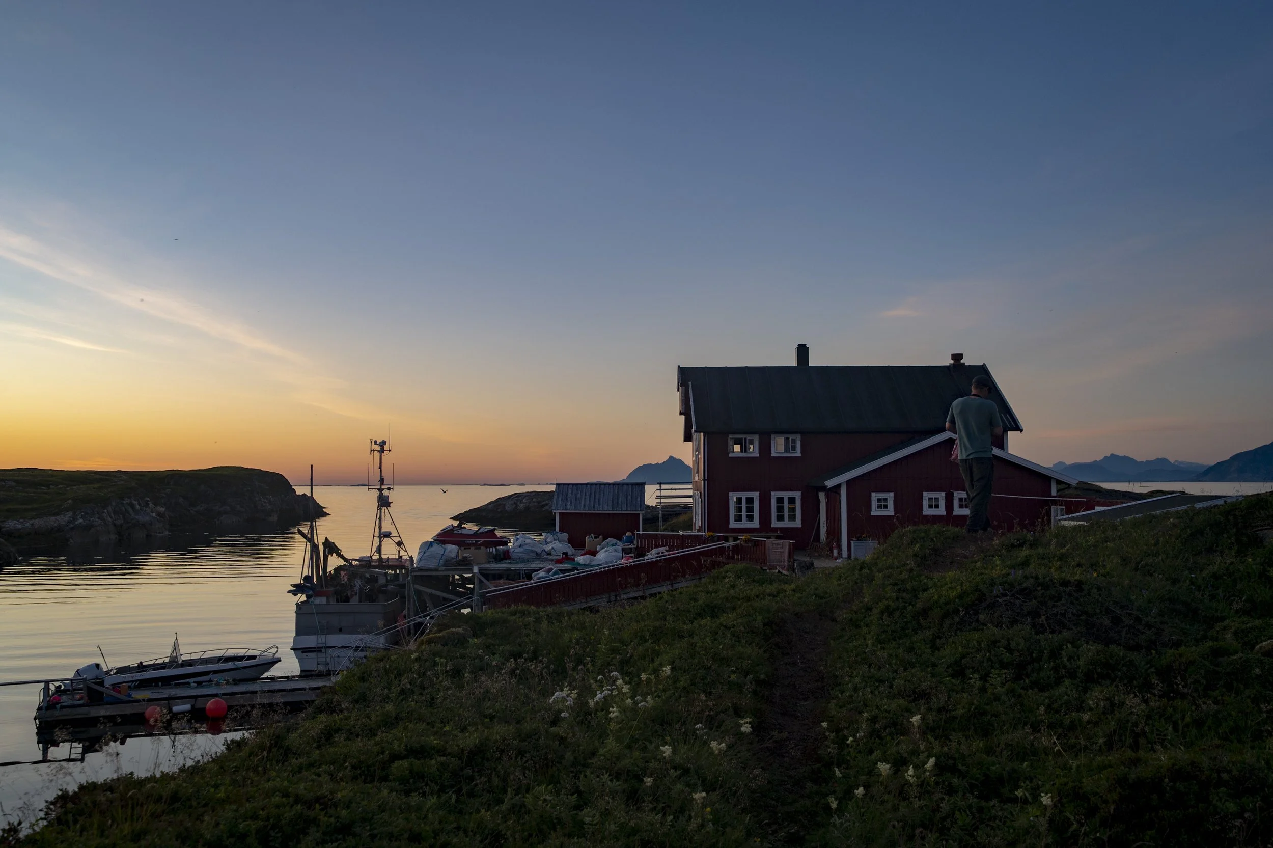 Et lite hus ved sjøen ved solnedgang, med en person som står på terrassen, båter i vannet og fjell i bakgrunnen.