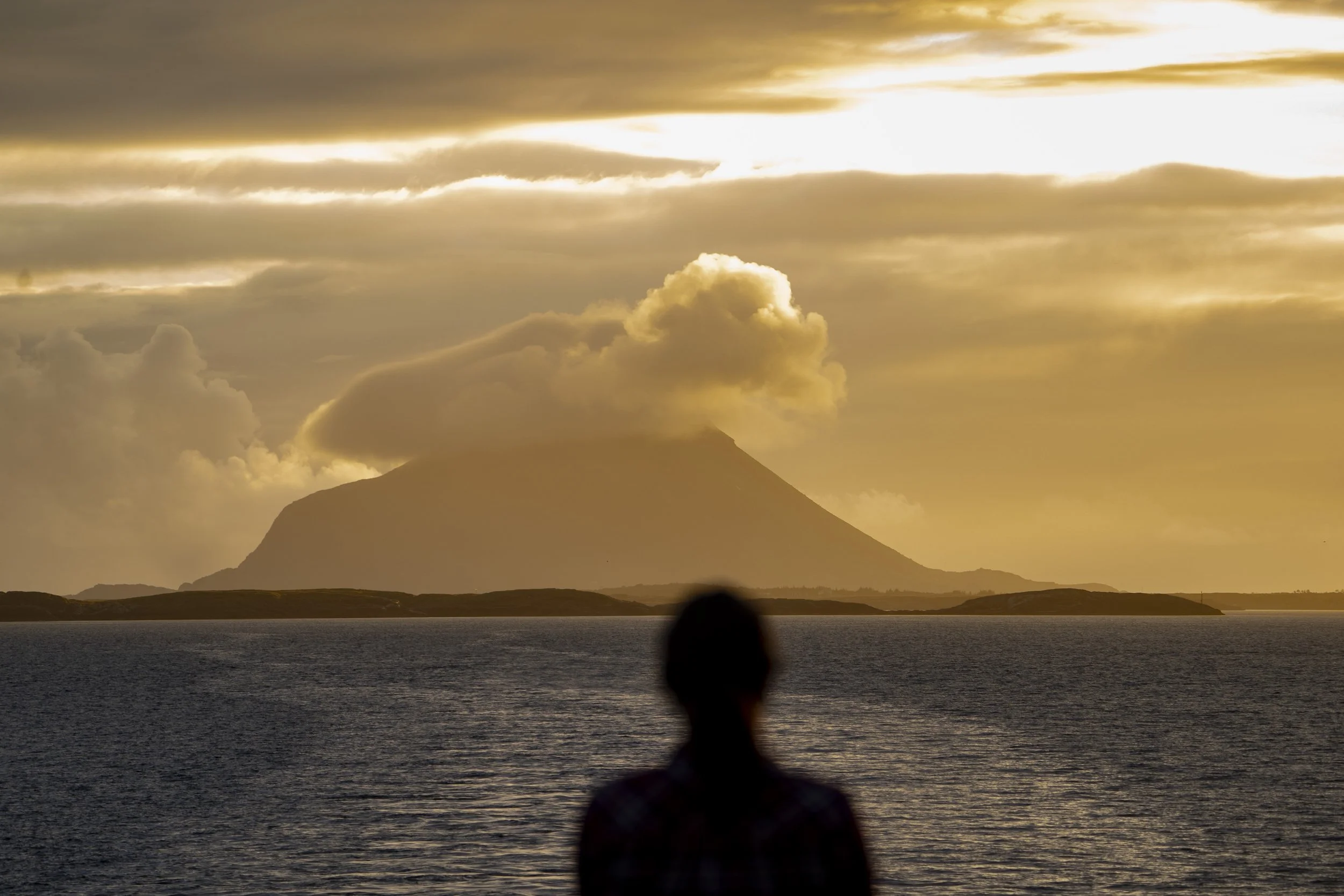 Silhuett av en person foran en storsjø med fjell i bakgrunnen, solnedgang med oransje og gylne himmelfarger, og en aktiv vulkan med røykskyer som stiger opp.