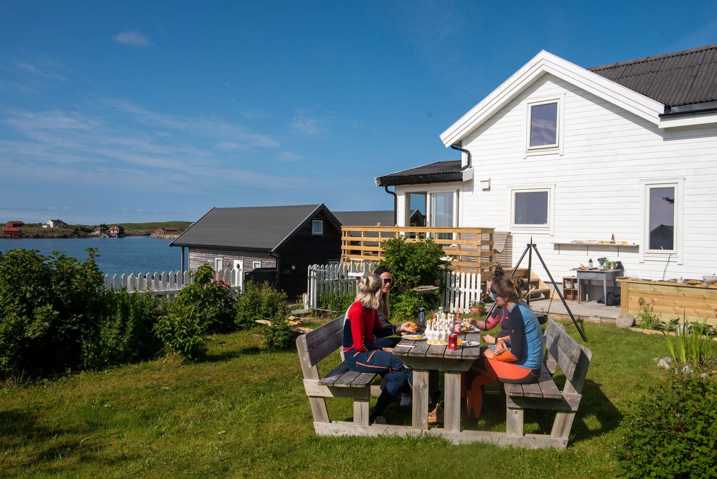 Folk som spiser ute i en hage med utsikt over sjøen, med hvite og mørke hus i bakgrunnen på en solrik dag.