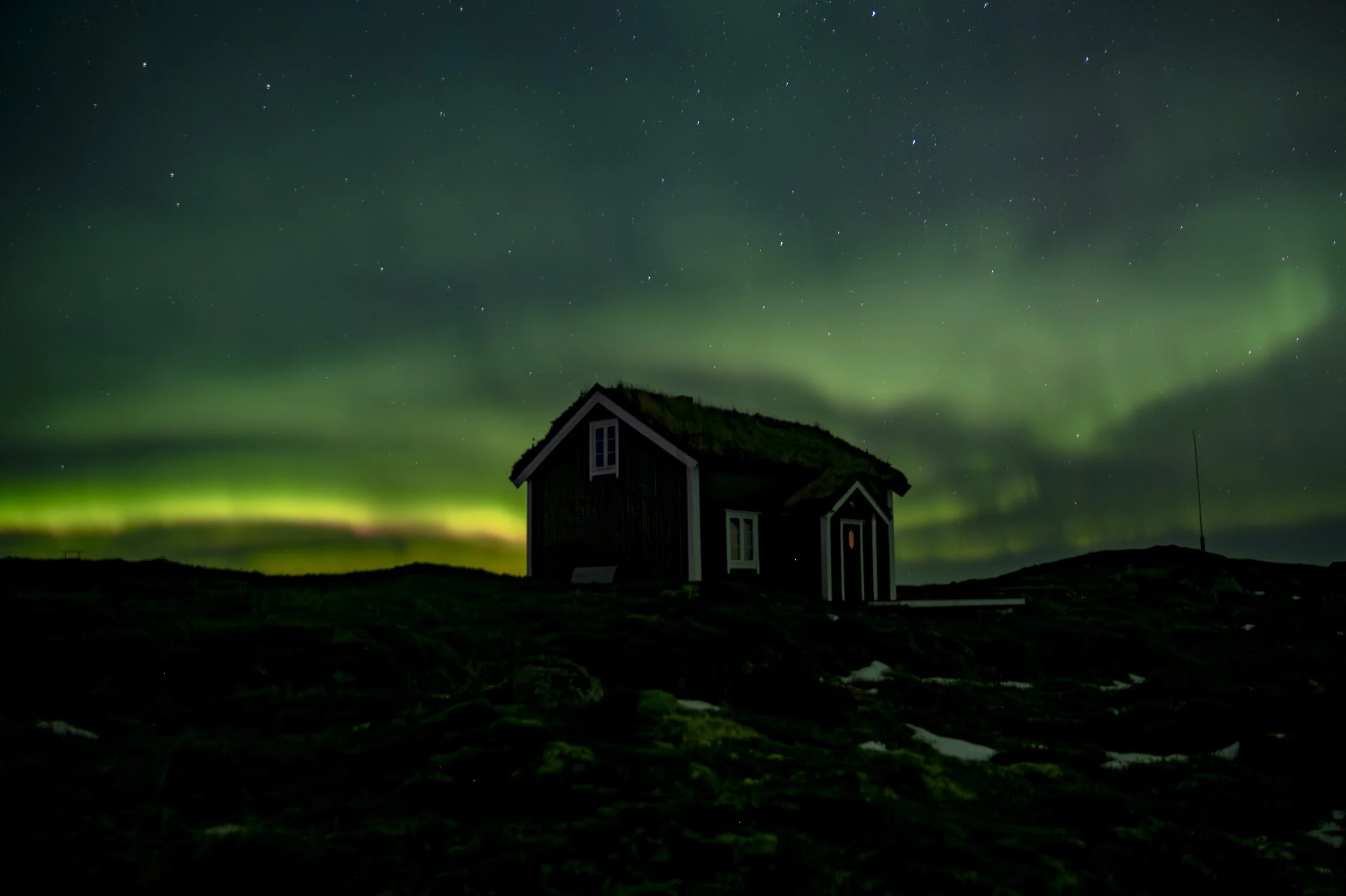 Nordlys over en liten hytte i mørket med en stjernefylt himmel.