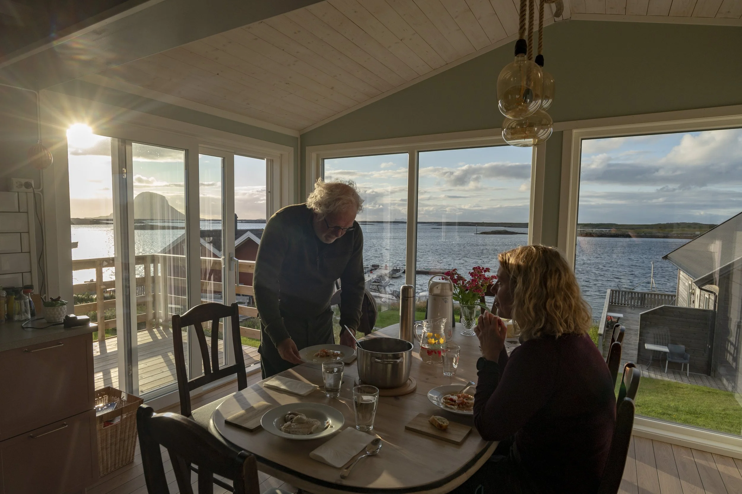 Interiør av et spisestue med store vinduer som viser utsikt over en sjø og en fjelltopp i solen. To personer, en eldre mann og en kvinne, spiser ved et rundt trebord med mat. Taket er kledd med lys trepanel, og det henger glasslamper over bordet.