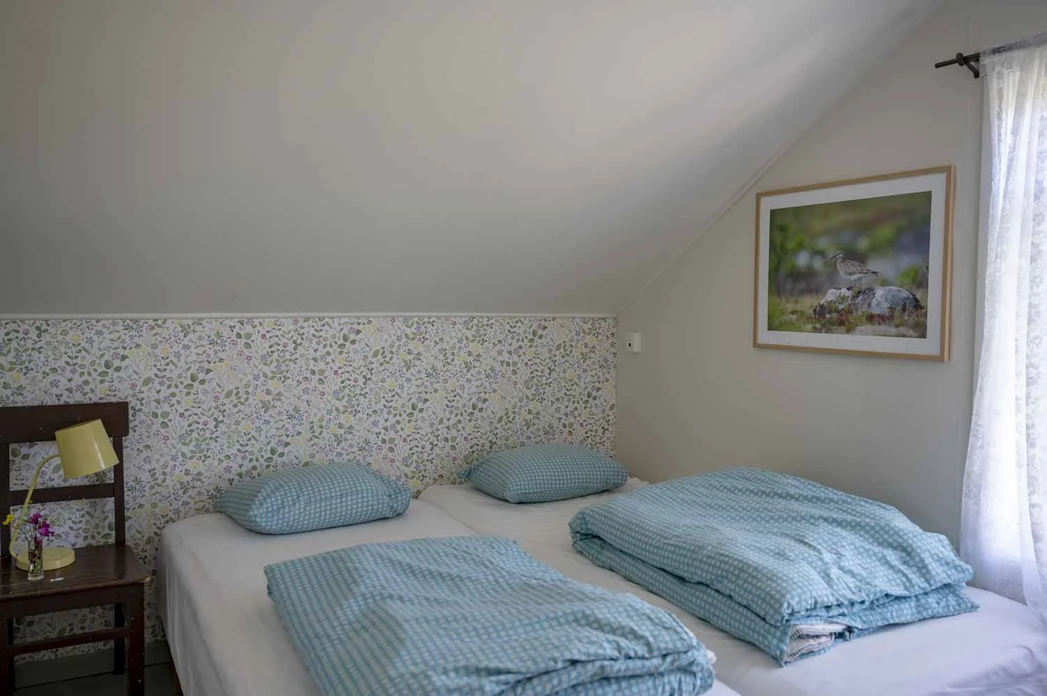 Soverom med seng, sengetøyet er blått med hvitt mønster, tapet bak sengen med blomster- og bladmotiver, en liten dagslampe på nattbord til venstre, bilde av en fugl på stein på veggen, lyst rom med vindu og gjennomsiktig gardin.