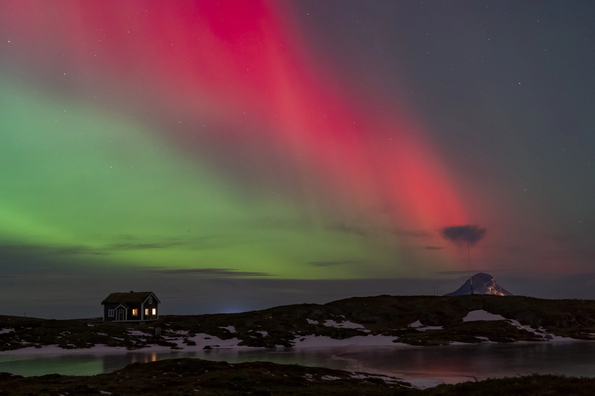 Fargede nordlys over et lite hus på en fjelltopp med snø på bakken.