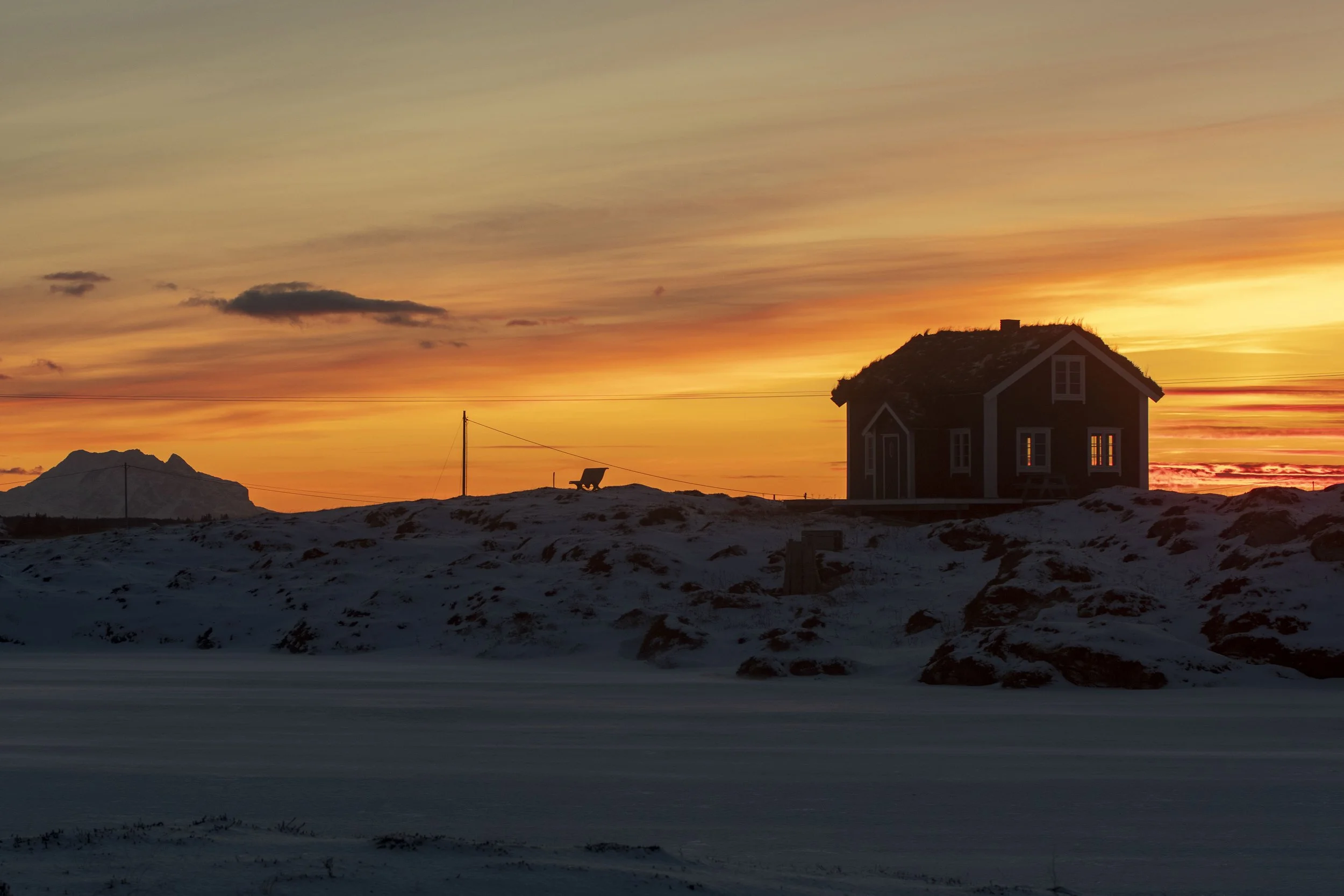Et lite hus med tak av gress står på en snødekt mark med en solnedgang i bakgrunnen. Det er også en lysbøyle av tråd med en hund som ligger på bakken i nærheten av huset.