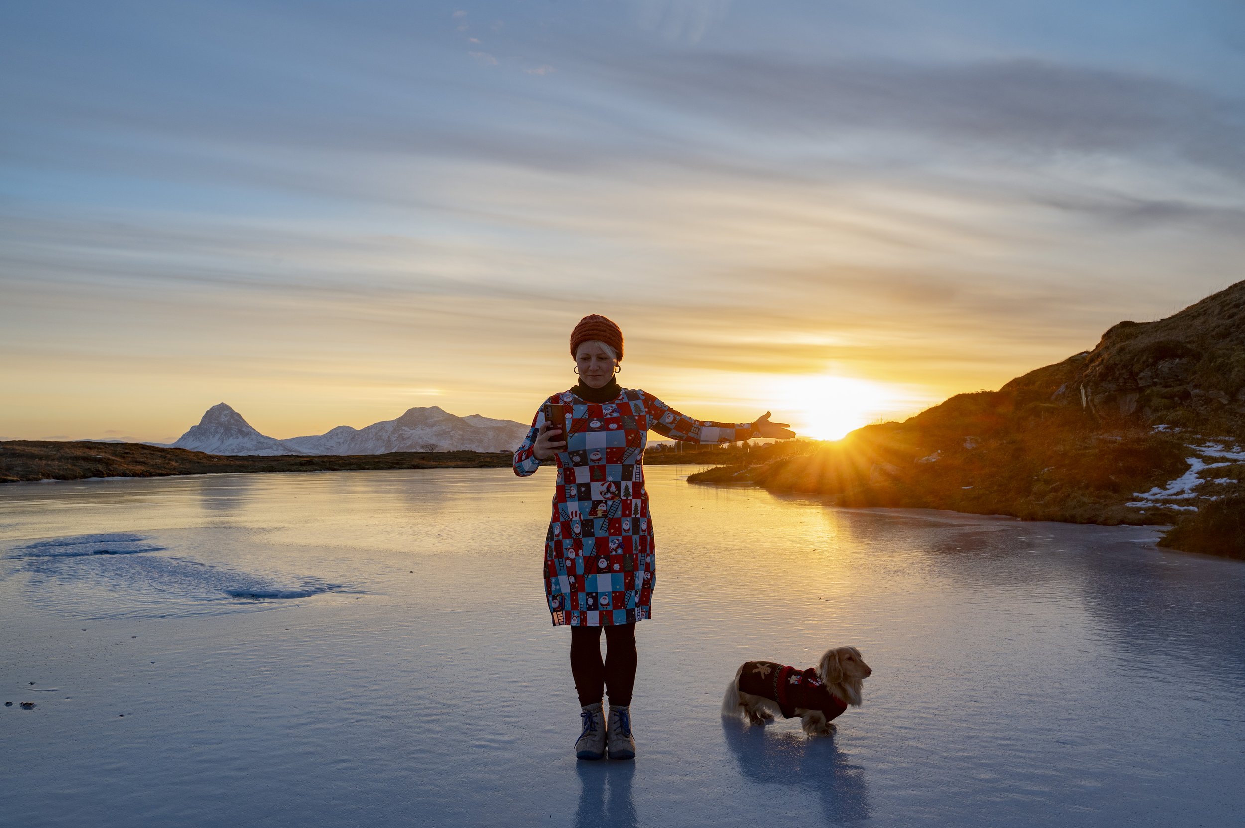 En kvinne med fargerik kjole og rød lue står ved en elv under solnedgangen, sammen med en hund.