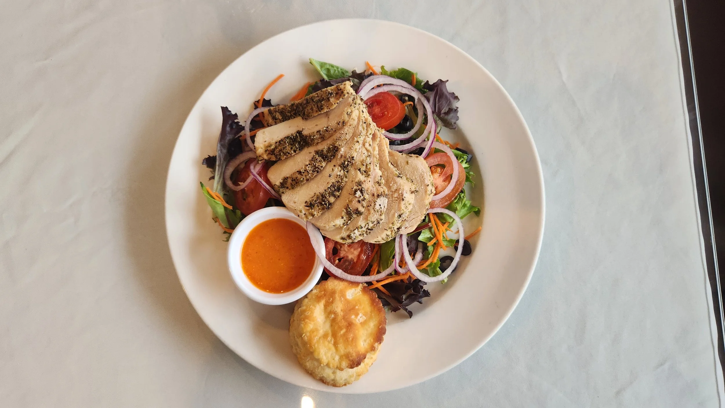 A photo of a green salad topped with sliced chicken breast, with dressing and a biscuit on the side.