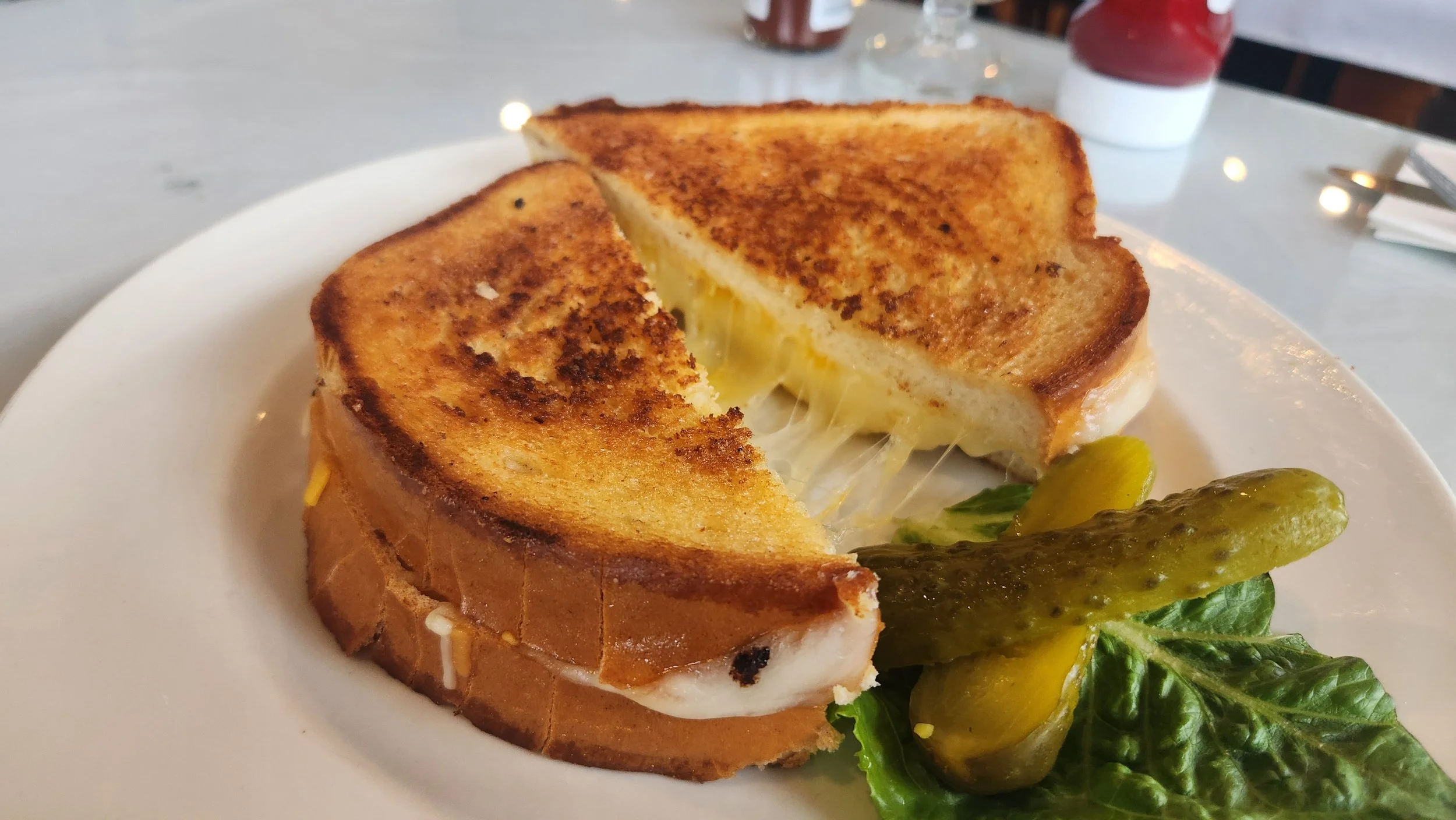 A photograph of a grilled cheese sandwich on sourdough bread, with a pickle on the side.