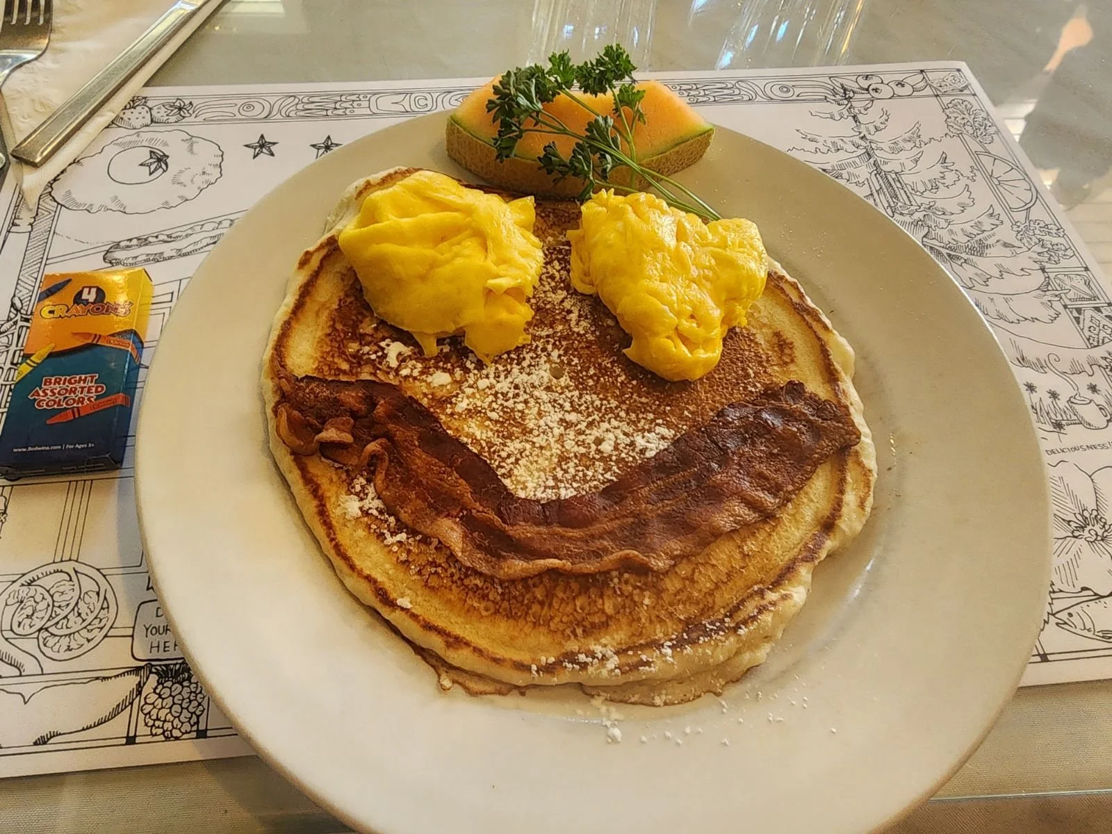 A photo of the Kid's breakfast combo plate with a pancake, scrambled eggs, and a slice of bacon arranged like a smiley face.
