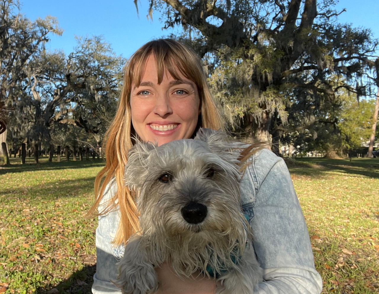 A woman smiling and holding a fluffy gray dog outdoors in a park with trees and blue sky in the background.
