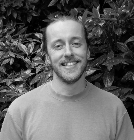 A smiling man with a beard and long hair tied back, standing outdoors in front of leafy plants.